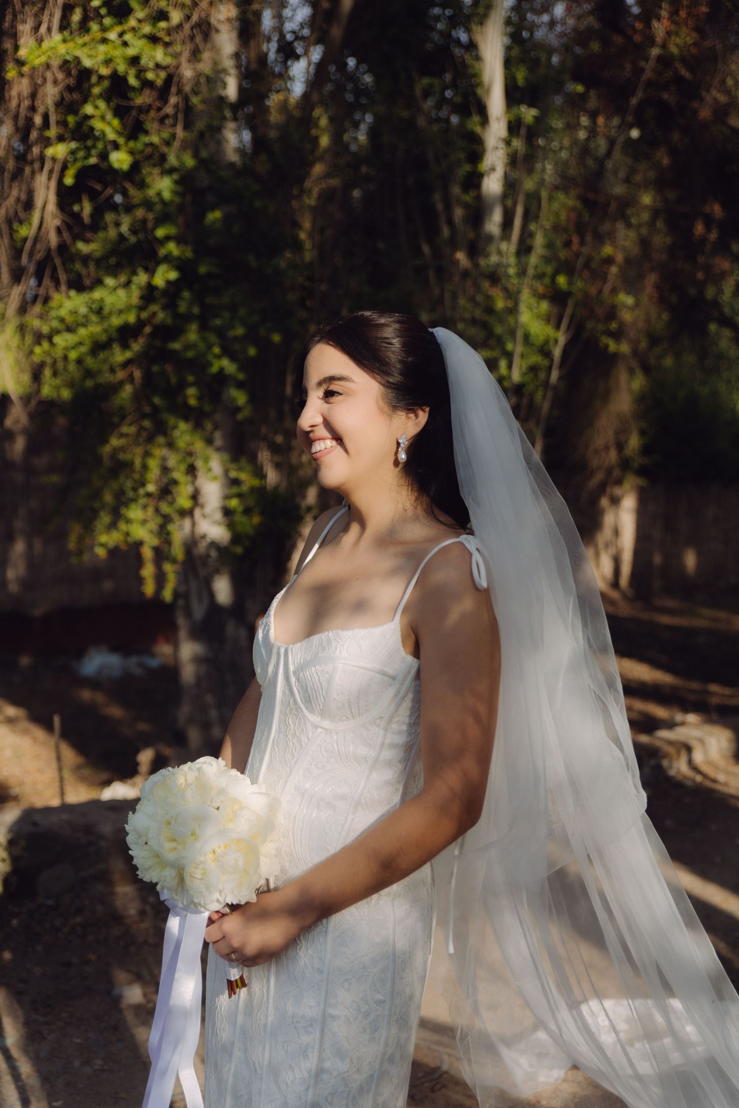 Elegant bride in white dress and veil gazes sideways while holding a white floral bouquet outdoors.