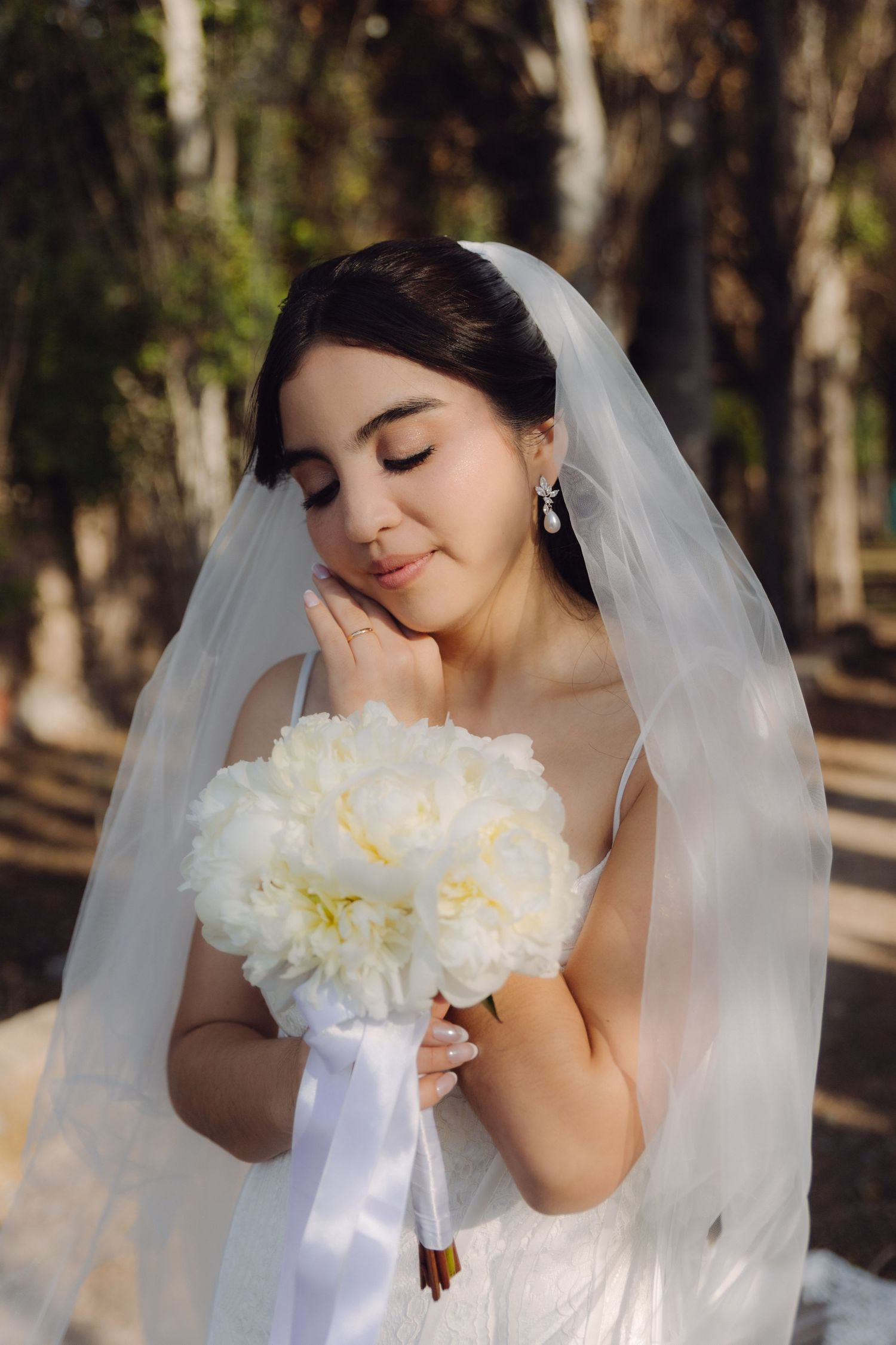 Serene bride with eyes closed holds white peony bouquet while wearing a veil in a sunny garden.