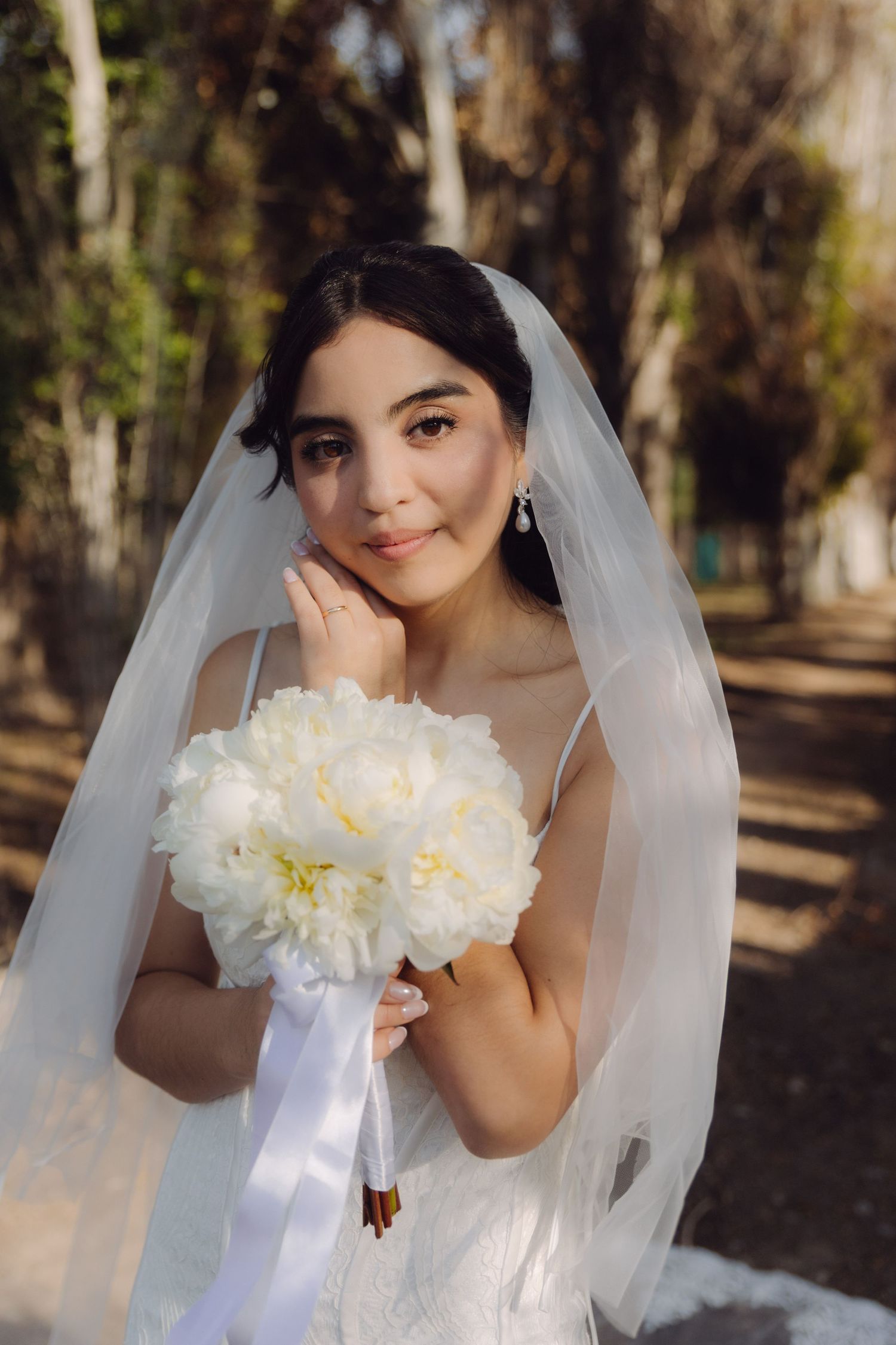Beautiful bride in white lace gown and veil poses with white peony bouquet in a sunlit park.
