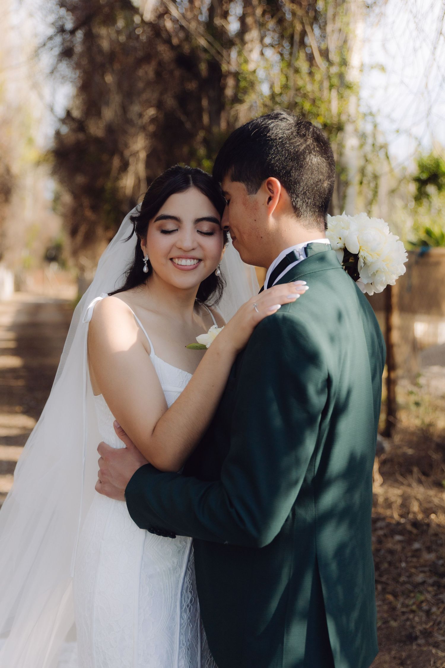 Happy bride smiles as groom embraces her tenderly in a sunlit outdoor wedding photo.