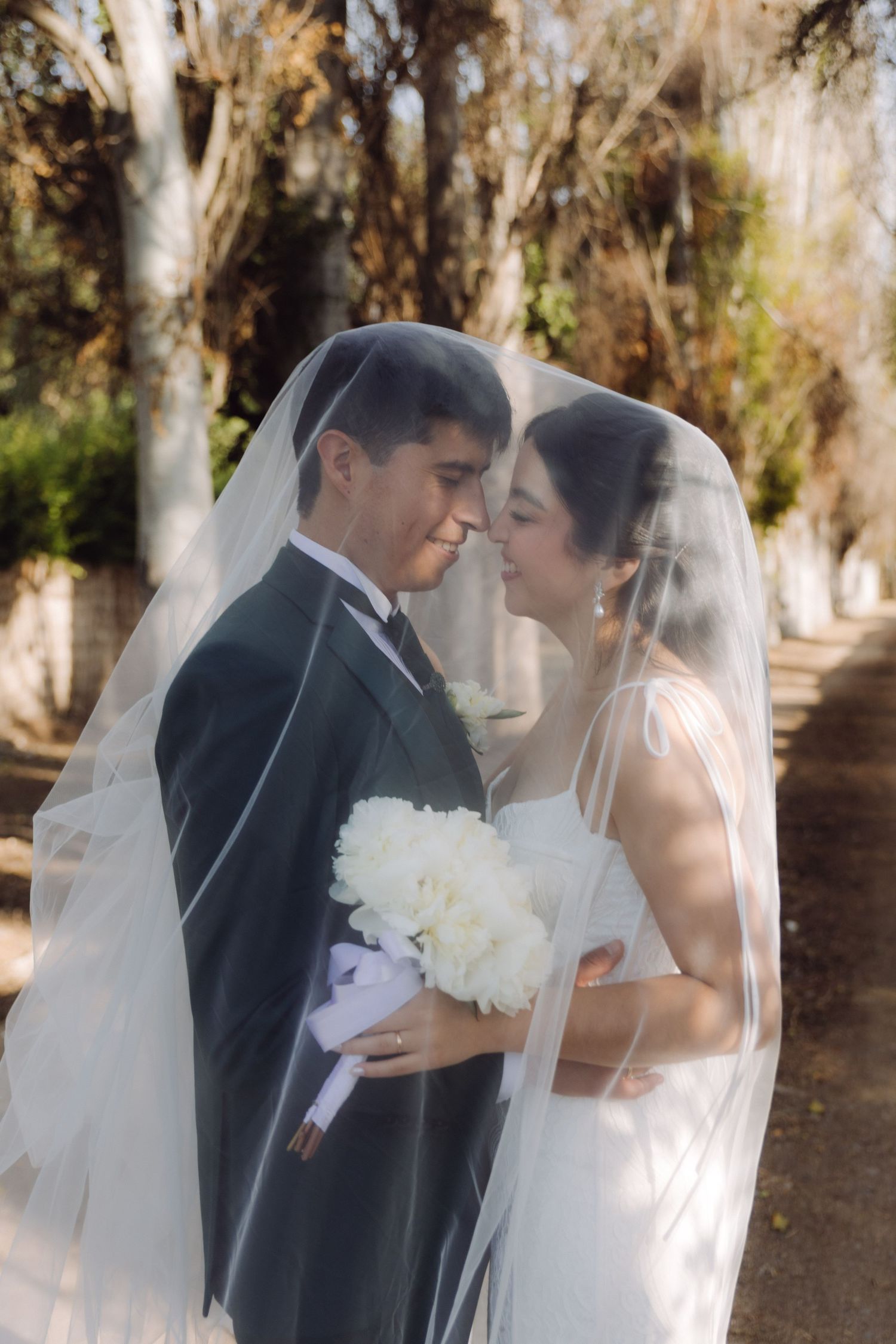 Bride and groom share an intimate moment under a white wedding veil outdoors, bride holding white peony bouquet.