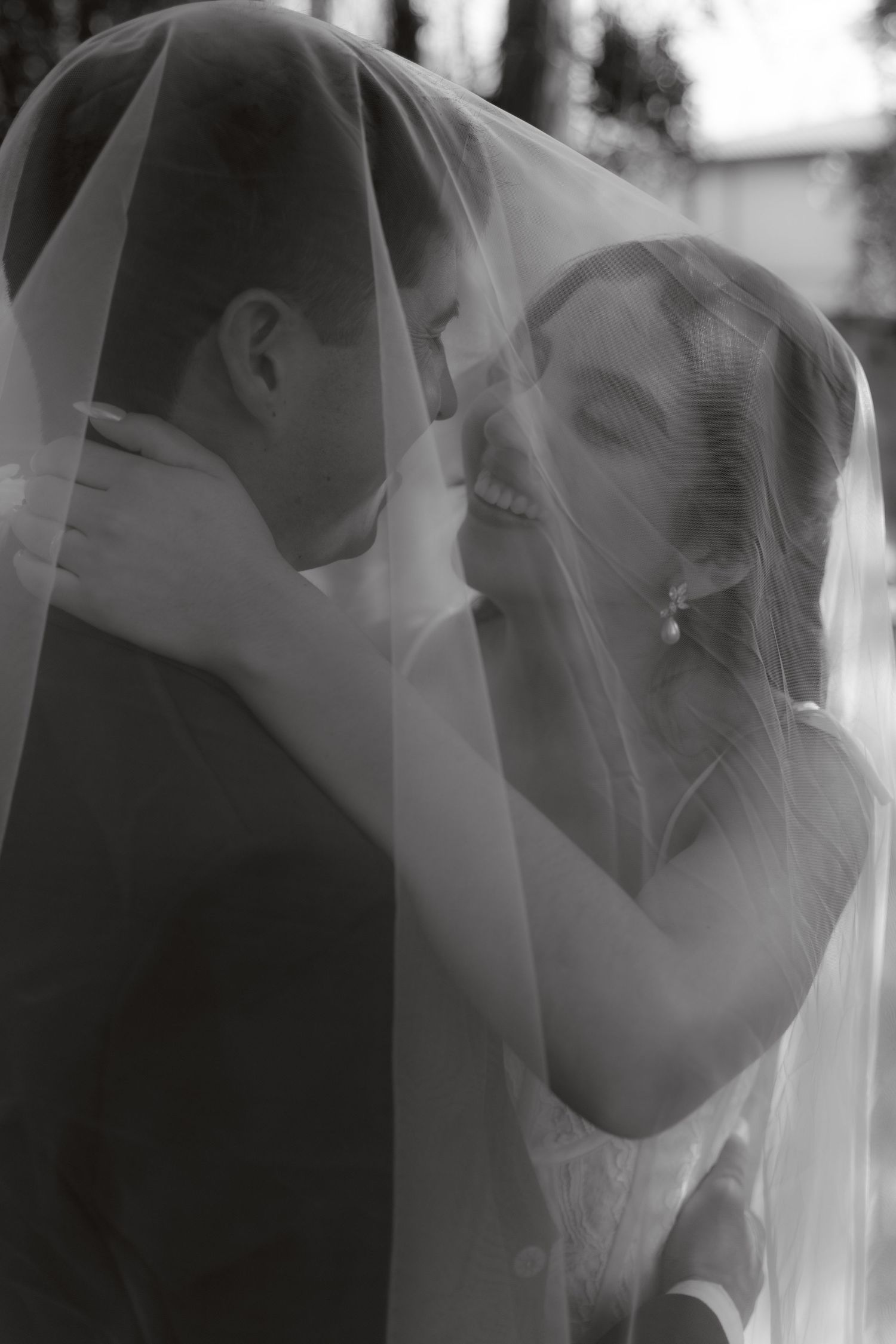 Black and white photo of smiling bride and groom embracing tenderly under a flowing wedding veil.
