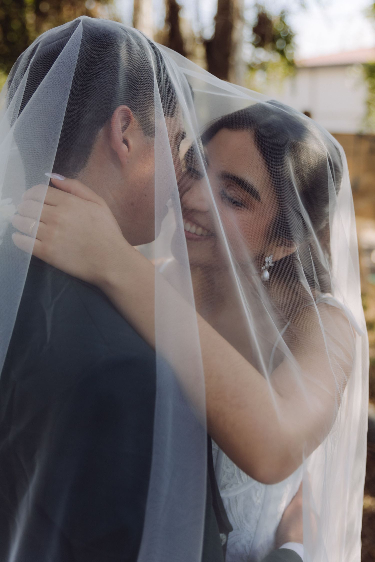 Joyful bride smiles while embracing groom under a sheer white wedding veil in outdoor setting.