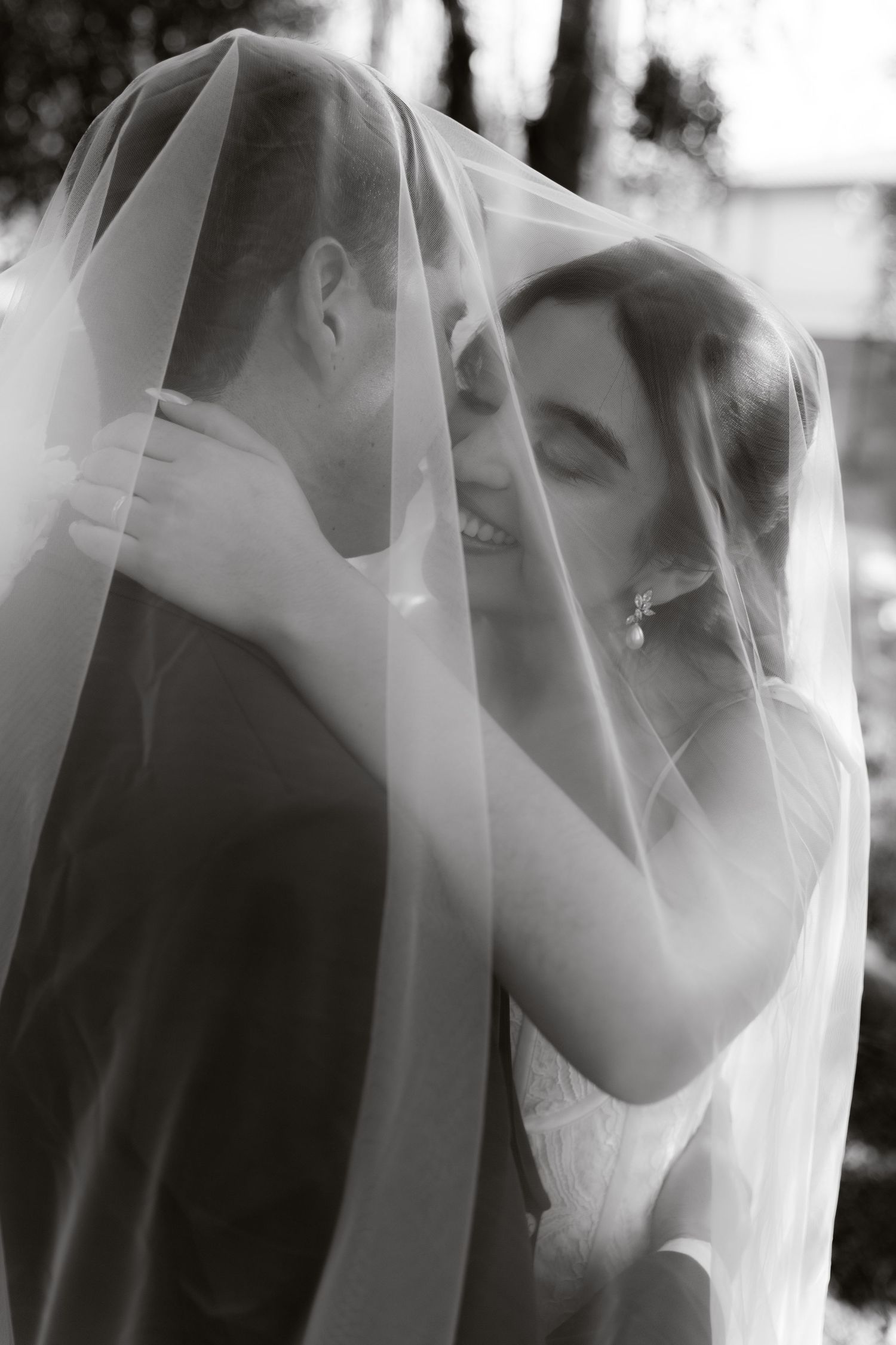 Black and white wedding photo of laughing bride and groom sharing romantic moment under bridal veil.