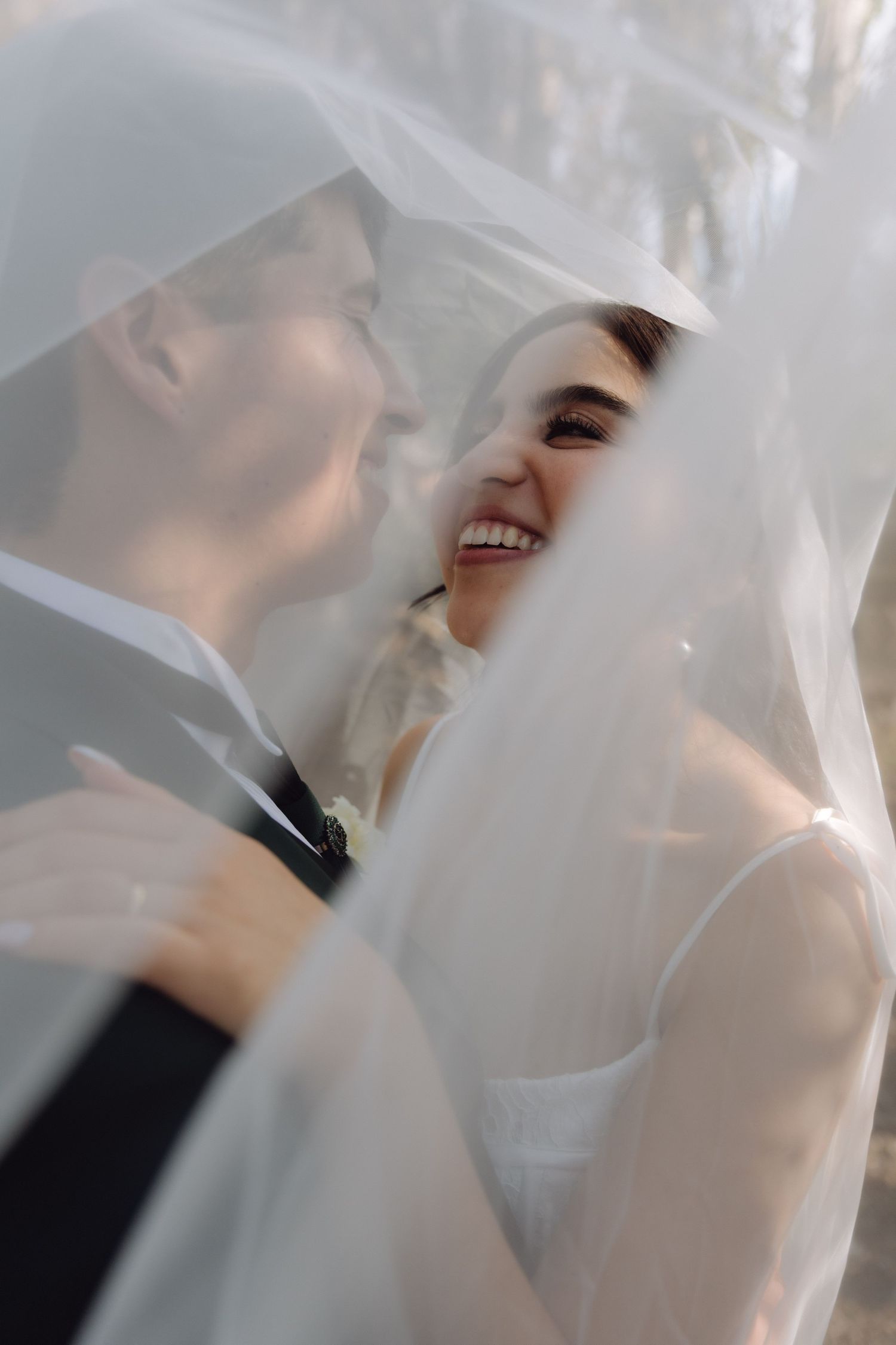 Happy bride laughs joyfully while standing close to groom beneath a draped white wedding veil.