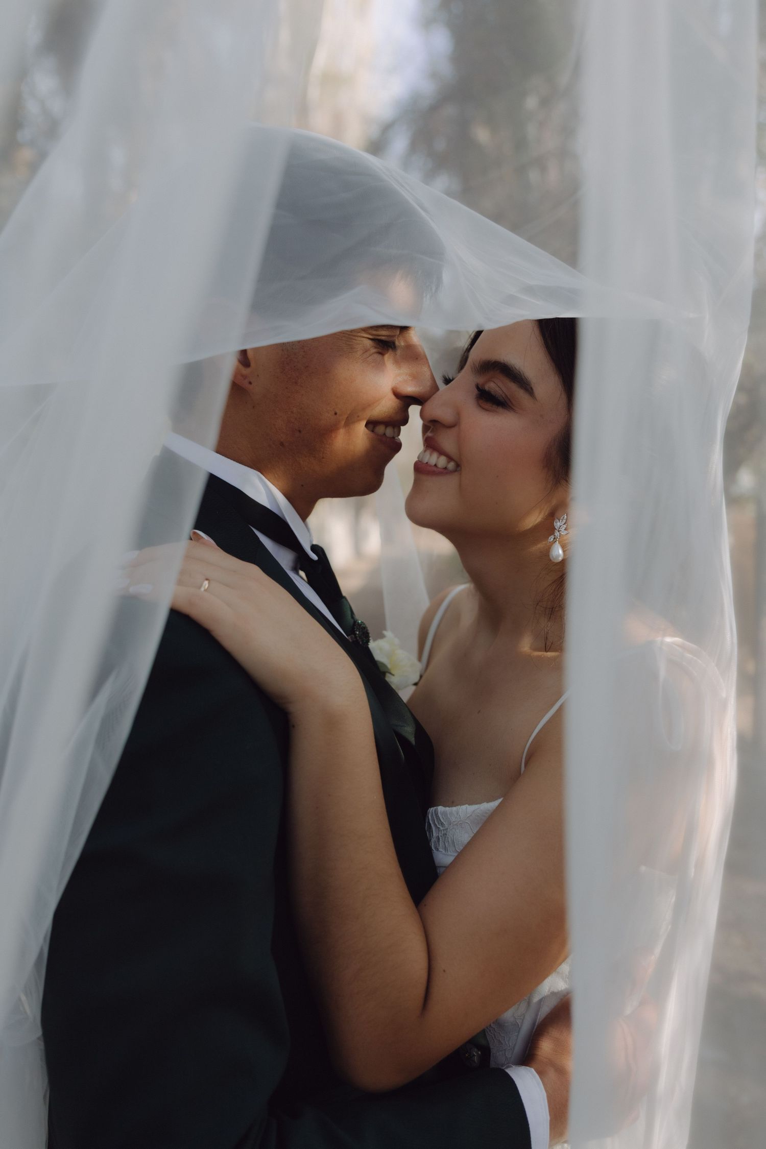Smiling bride and groom nearly kissing framed by flowing white veil in soft natural light.
