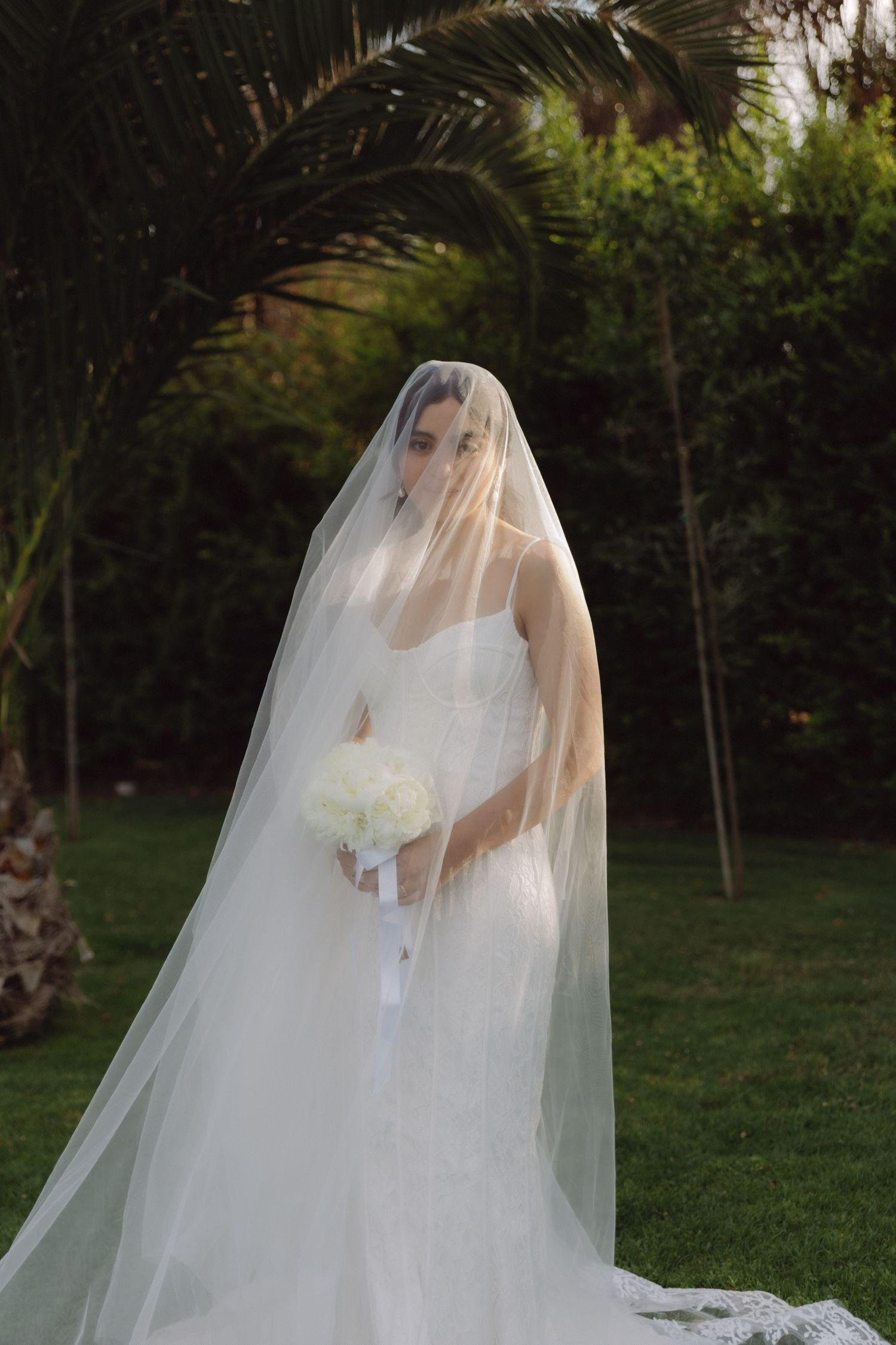 Elegant bride in white gown and long veil covering face holds white bouquet near palm trees.