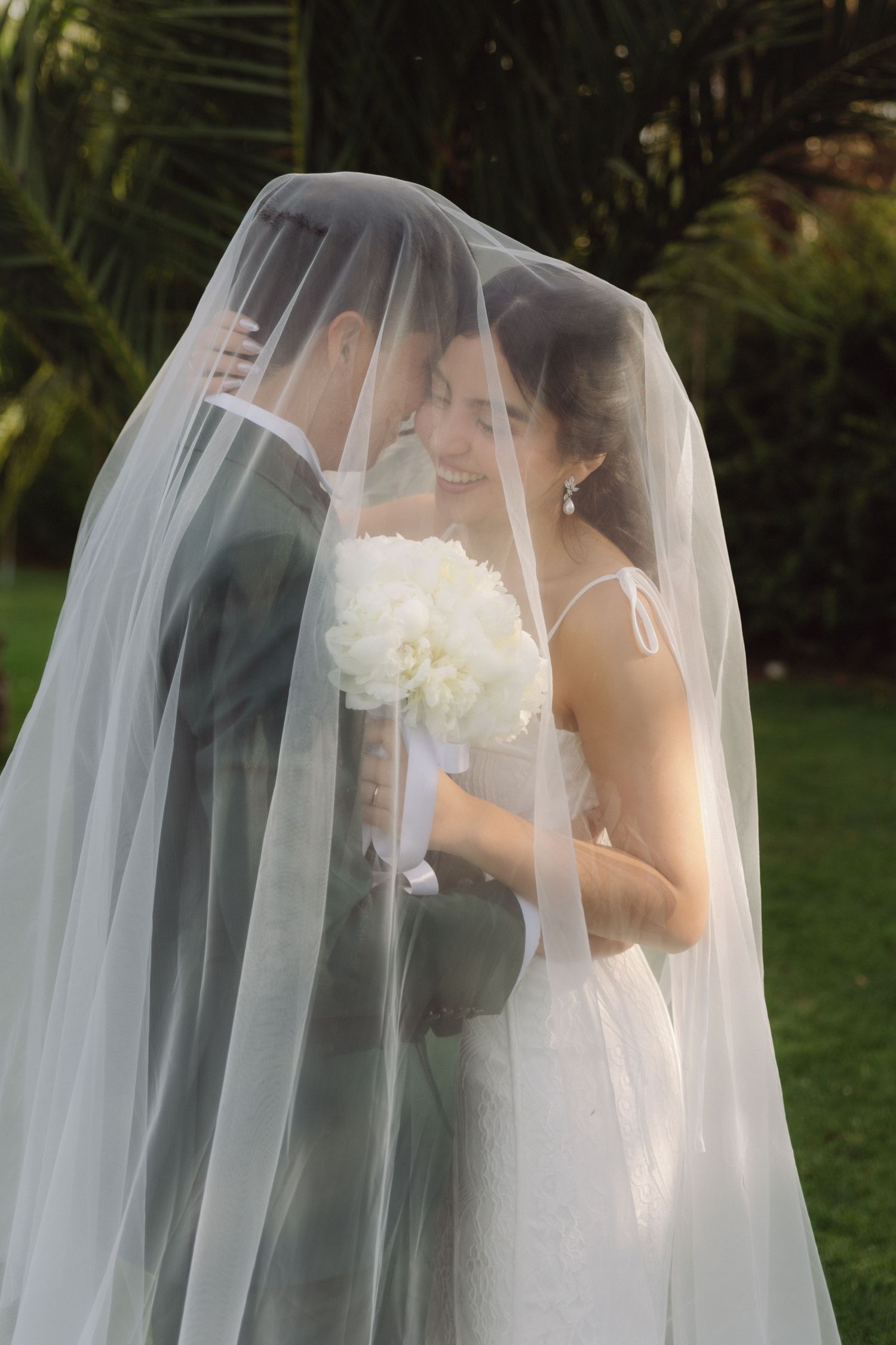 Wedding couple nearly kissing beneath sheer bridal veil with lush green palm trees in background.