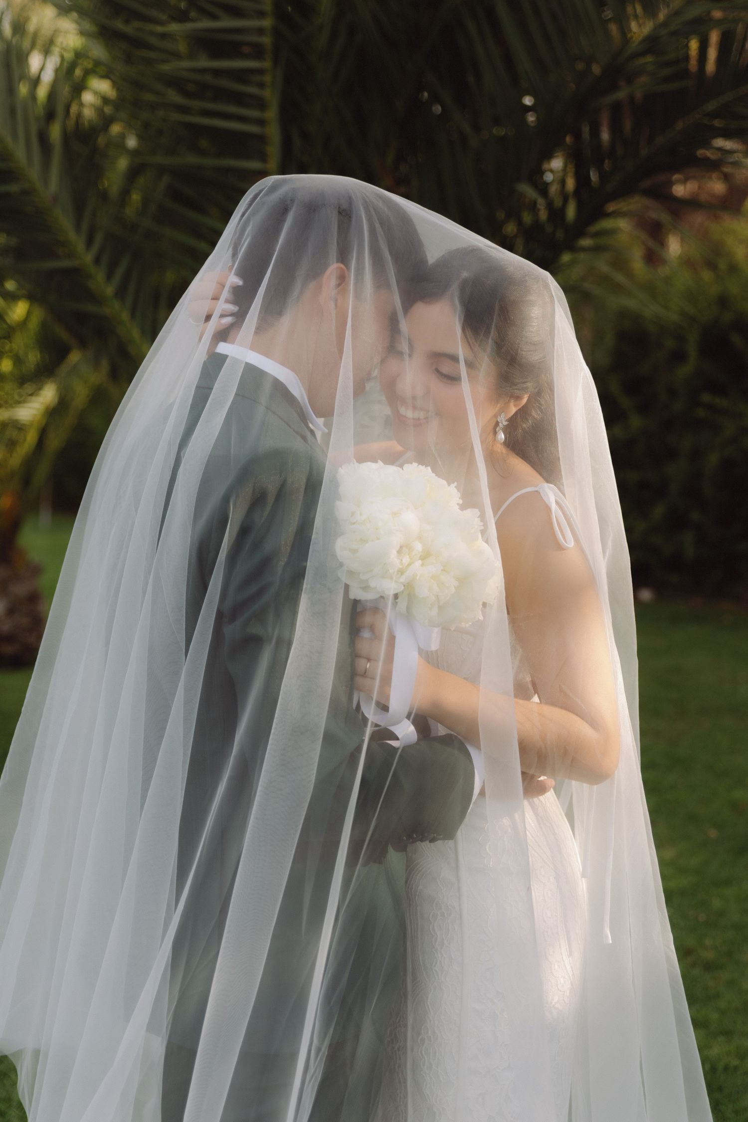 Bride and groom share intimate moment under flowing white wedding veil outdoors, bride holding white floral bouquet.