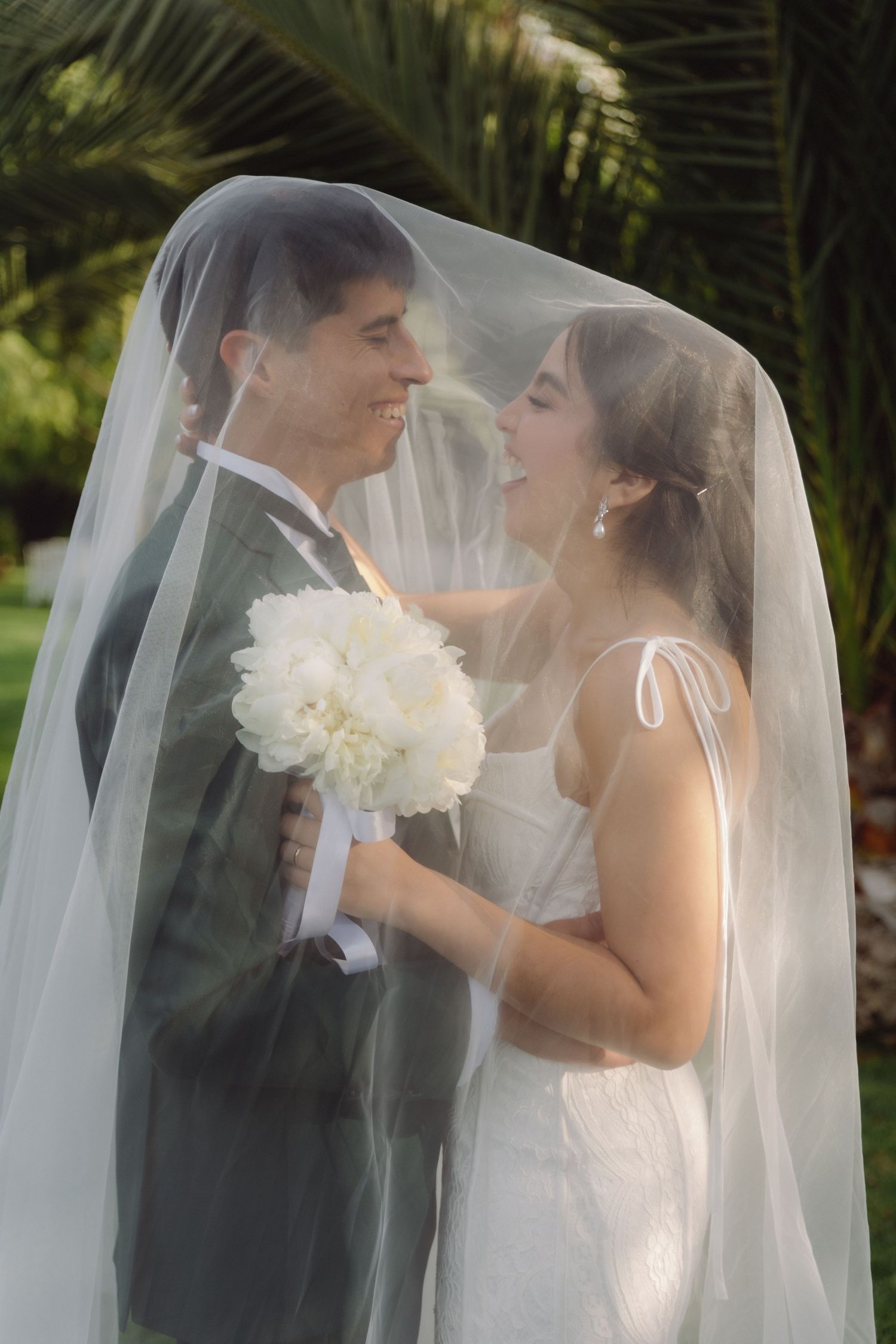 Smiling bride and groom share intimate moment under wedding veil outdoors.
