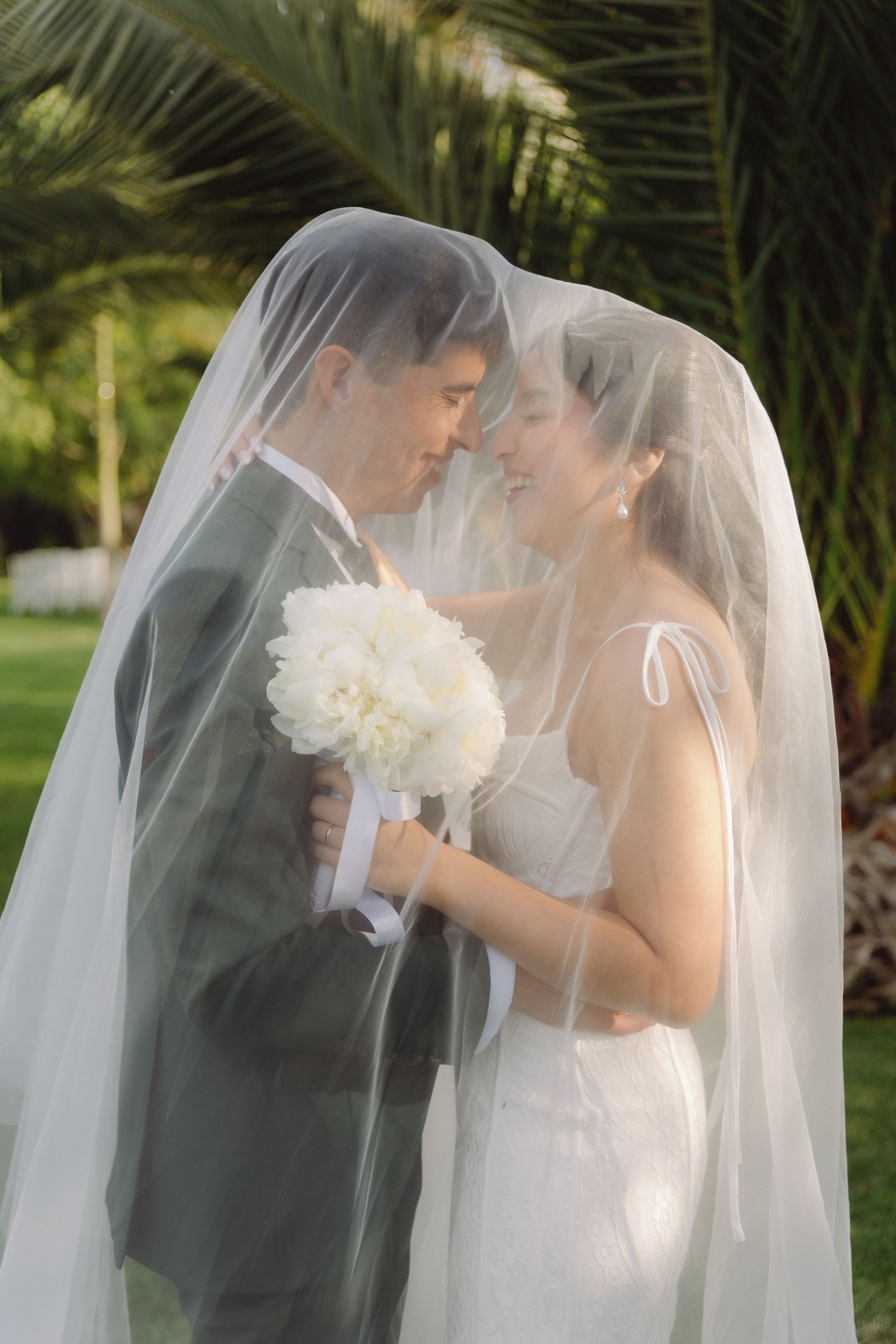 Happy bride and groom nearly kissing under sheer veil in garden.