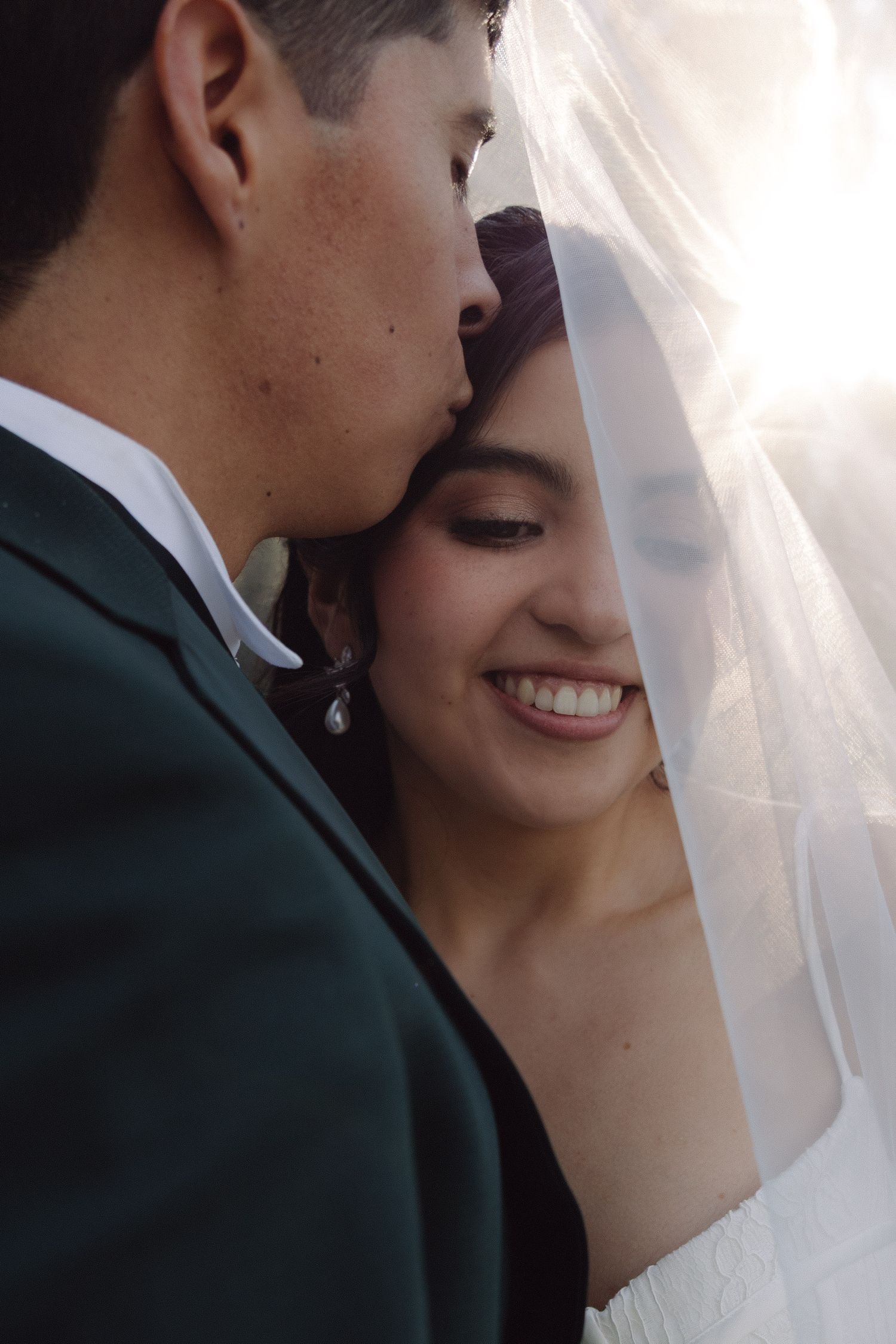 Groom kissing smiling bride on forehead under wedding veil in sunlight.