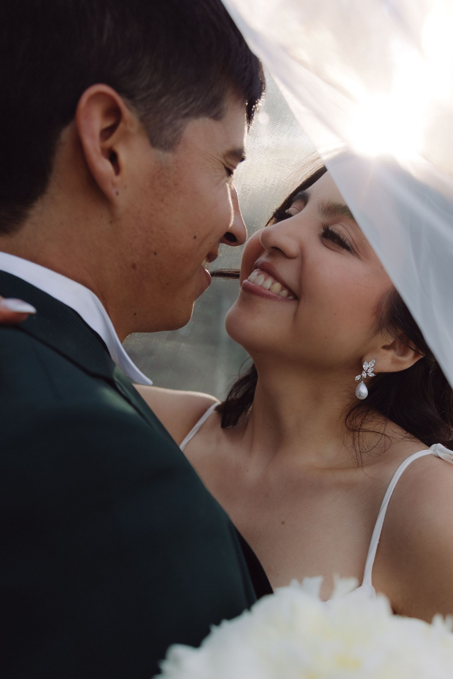 Close-up of smiling bride and groom nearly kissing under white veil.