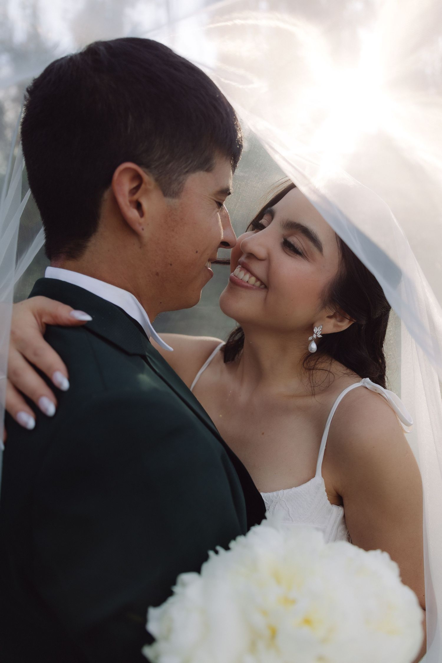 Joyful bride embracing groom under flowing veil with white bouquet outdoors.