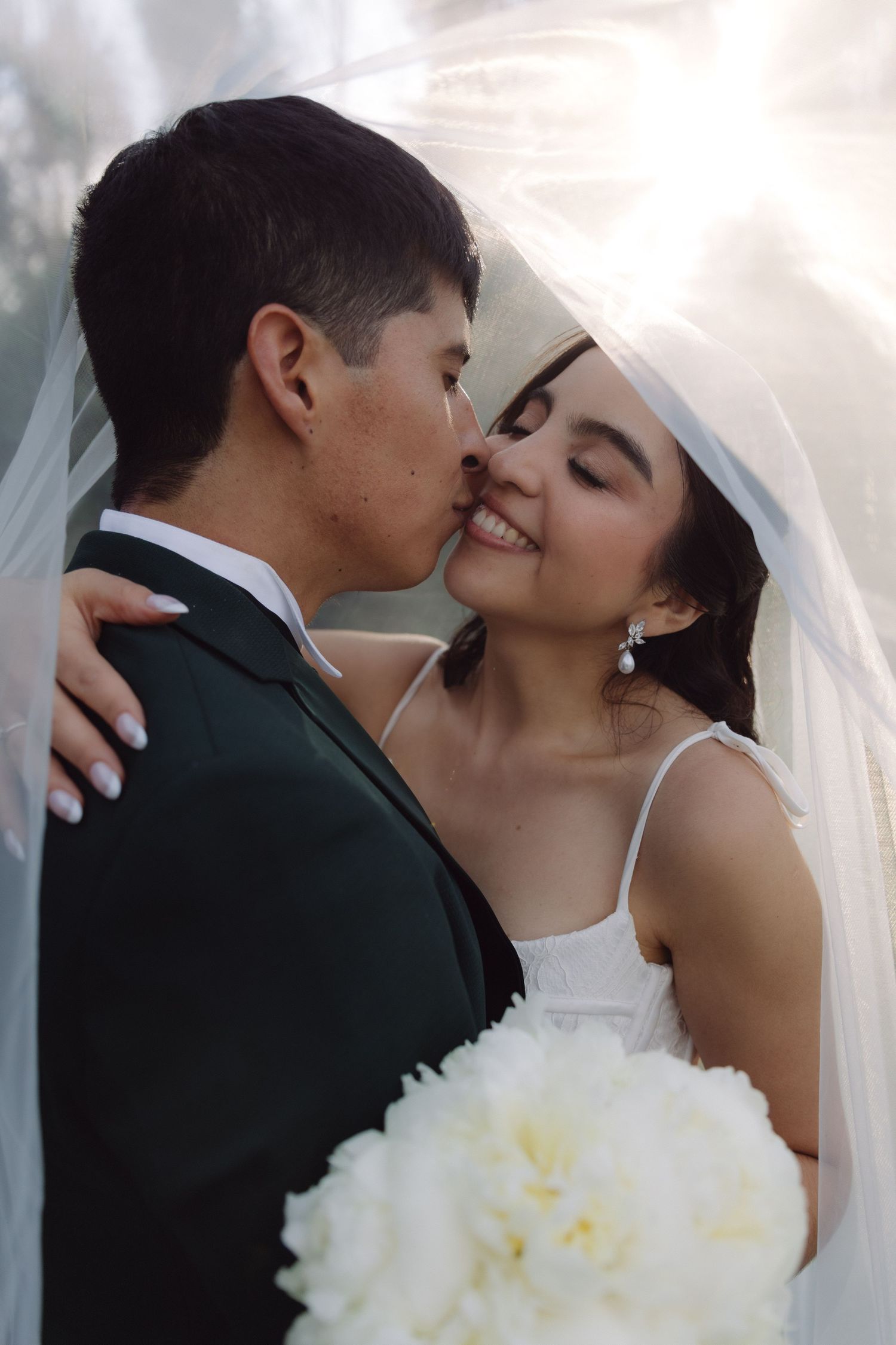 Bride and groom sharing a kiss under veil with white bouquet.