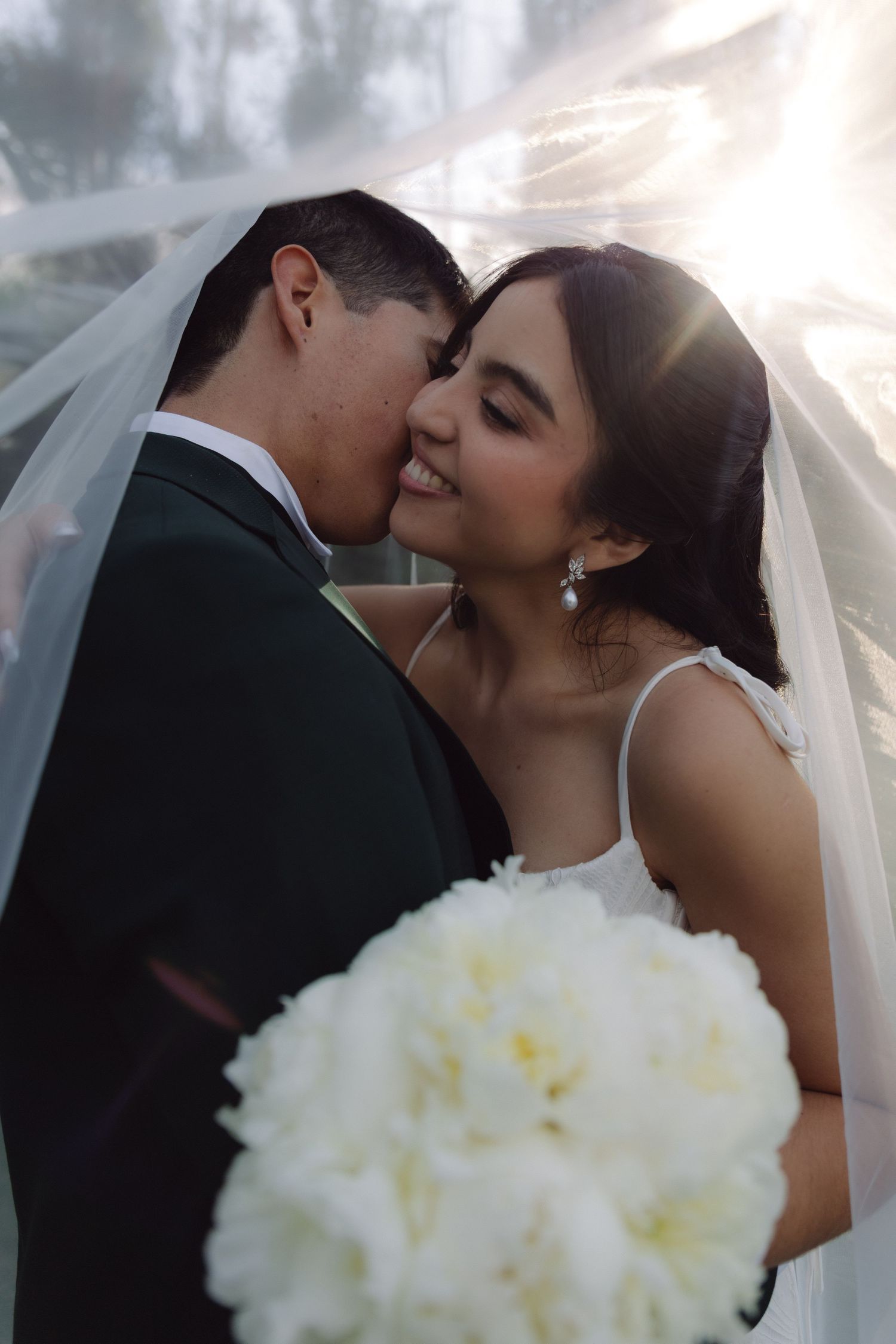 Romantic bride and groom kissing under wedding veil in golden sunlight.