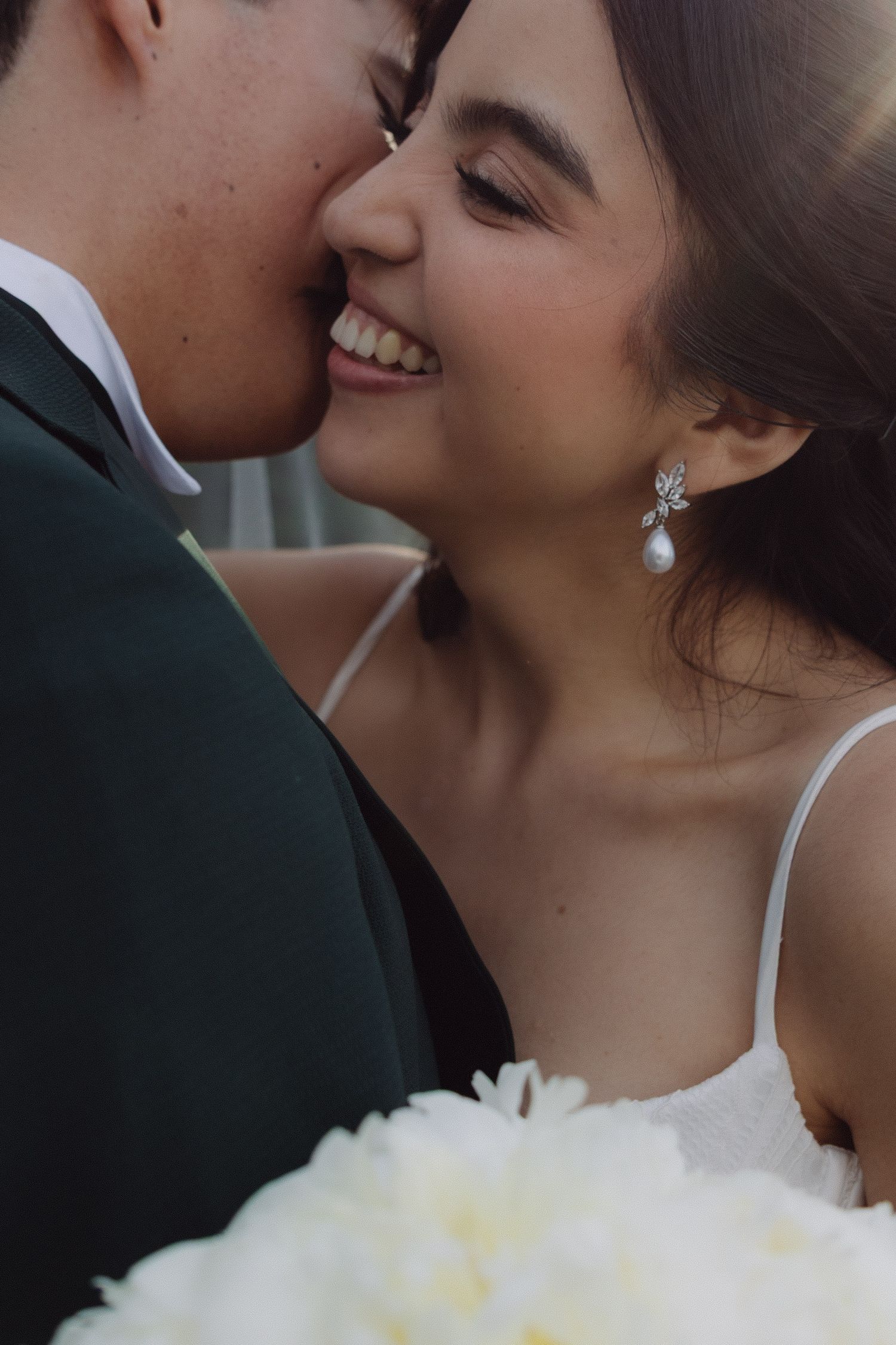 Close-up of laughing bride with pearl earrings beside groom holding bouquet.