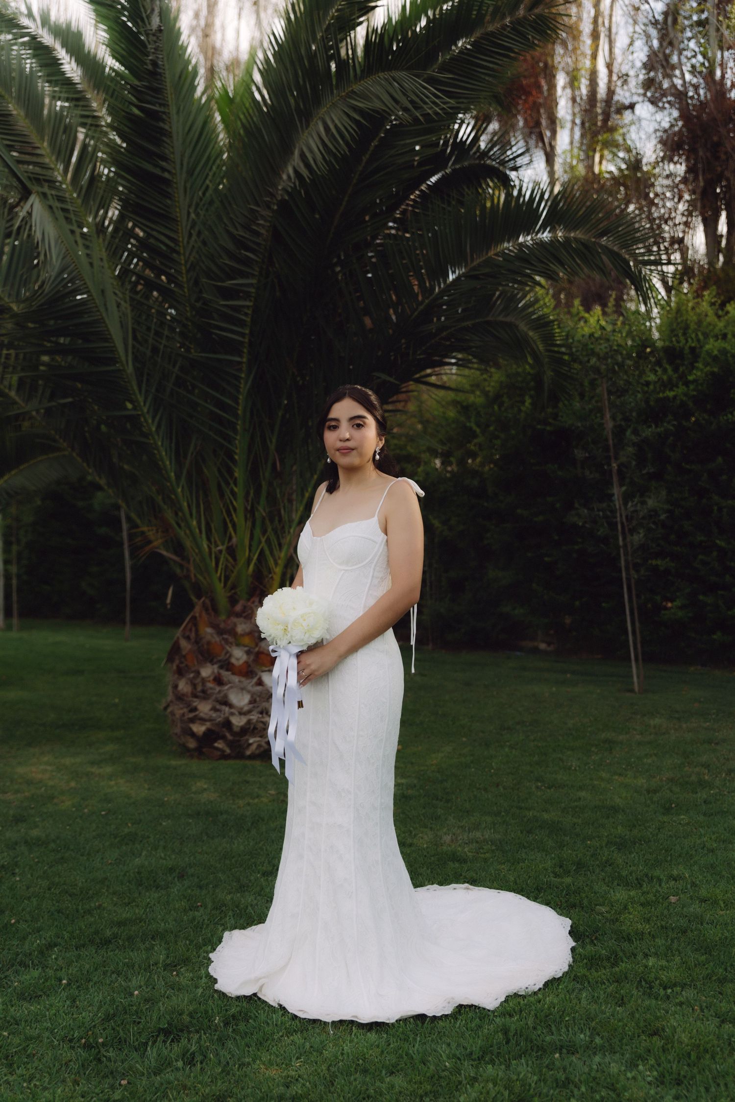 Elegant bride in fitted white gown holding bouquet beside tropical palm tree.