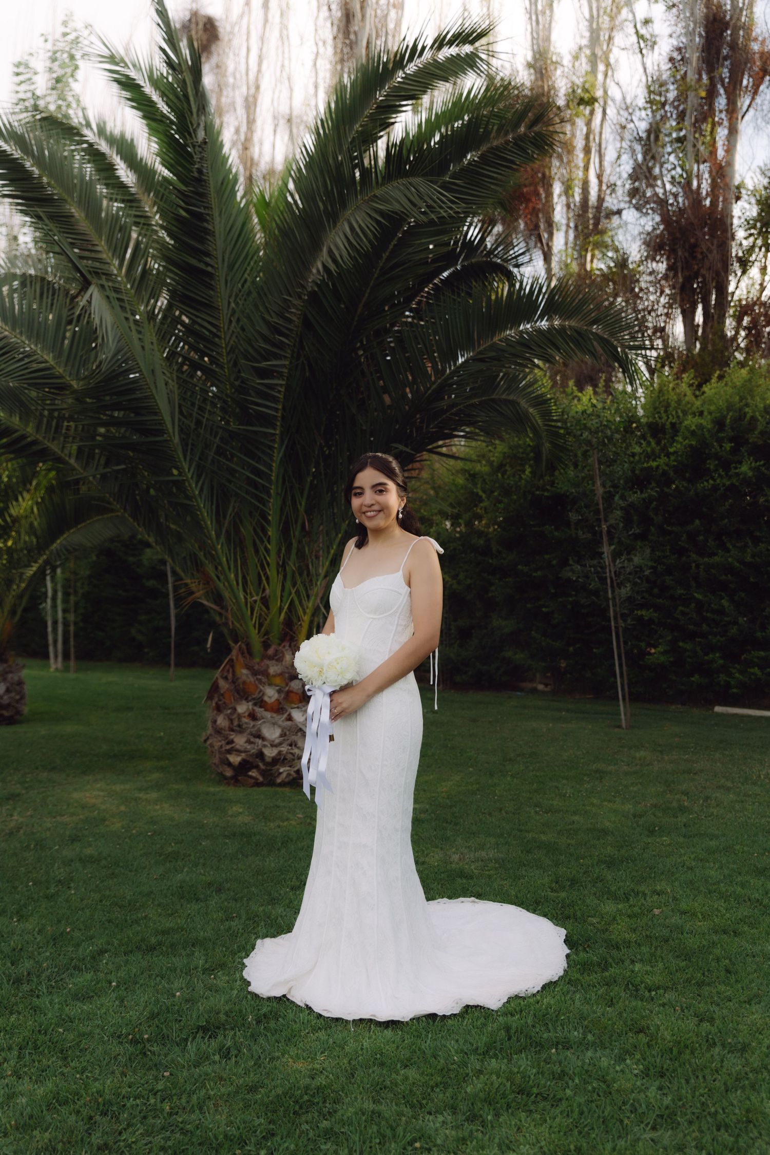 Bride in white mermaid gown holding bouquet standing before palm tree on green lawn.