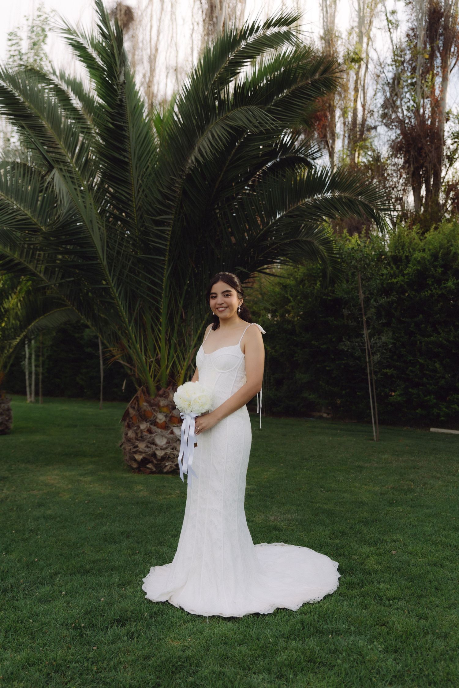 Elegant bride in fitted white dress with train poses with white bouquet by palm tree.