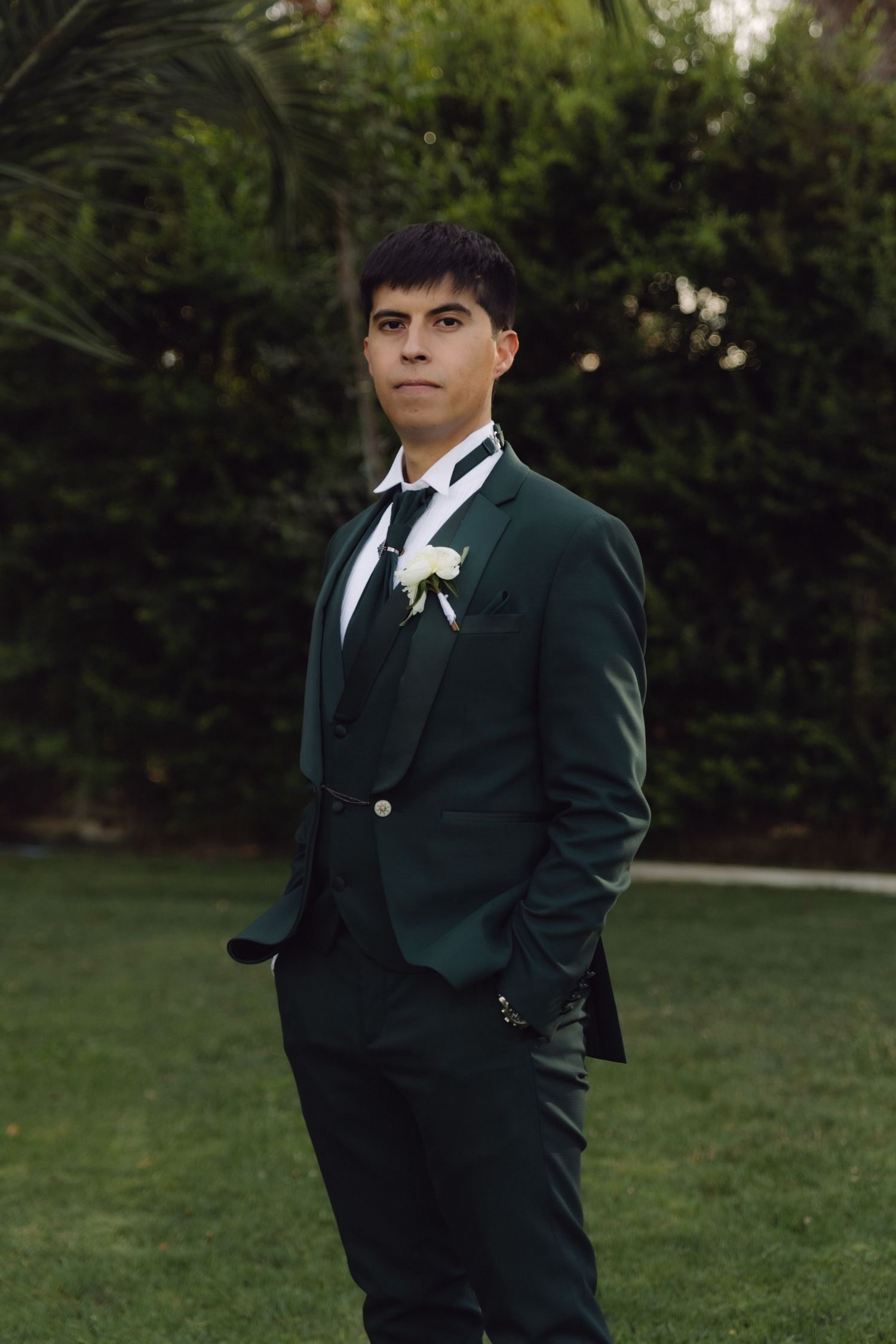 Groom in dark green three-piece suit with white boutonniere stands on garden lawn.
