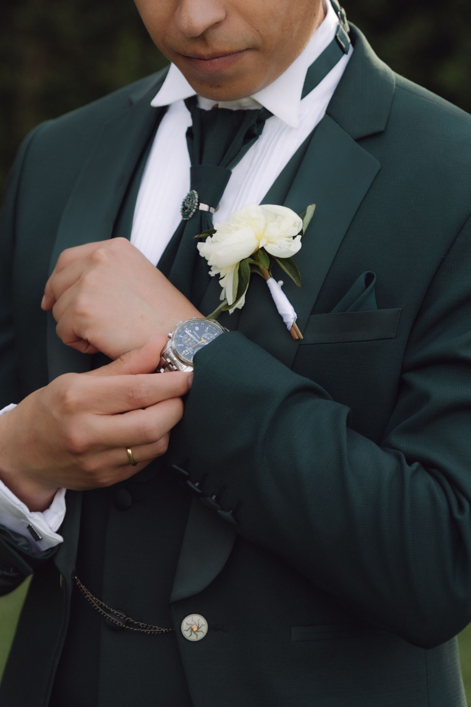 Close-up of groom in green suit adjusting watch cufflink with white peony boutonniere.