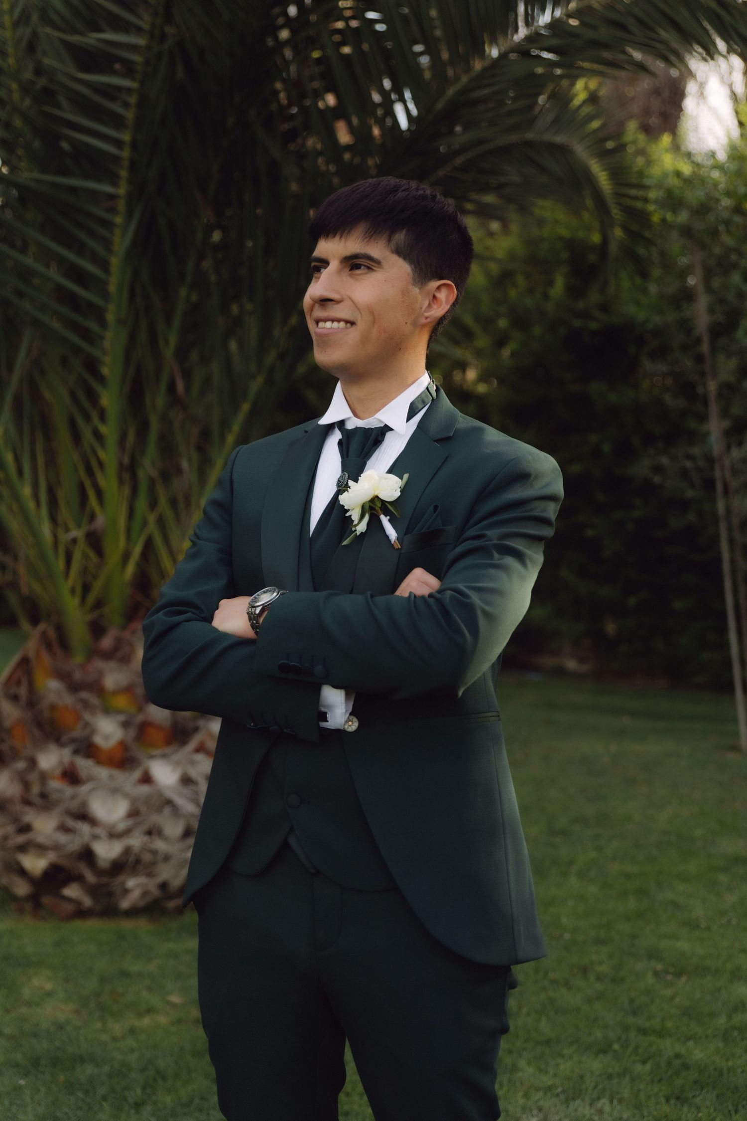 Smiling groom in dark green suit with arms crossed stands before tropical palm trees.