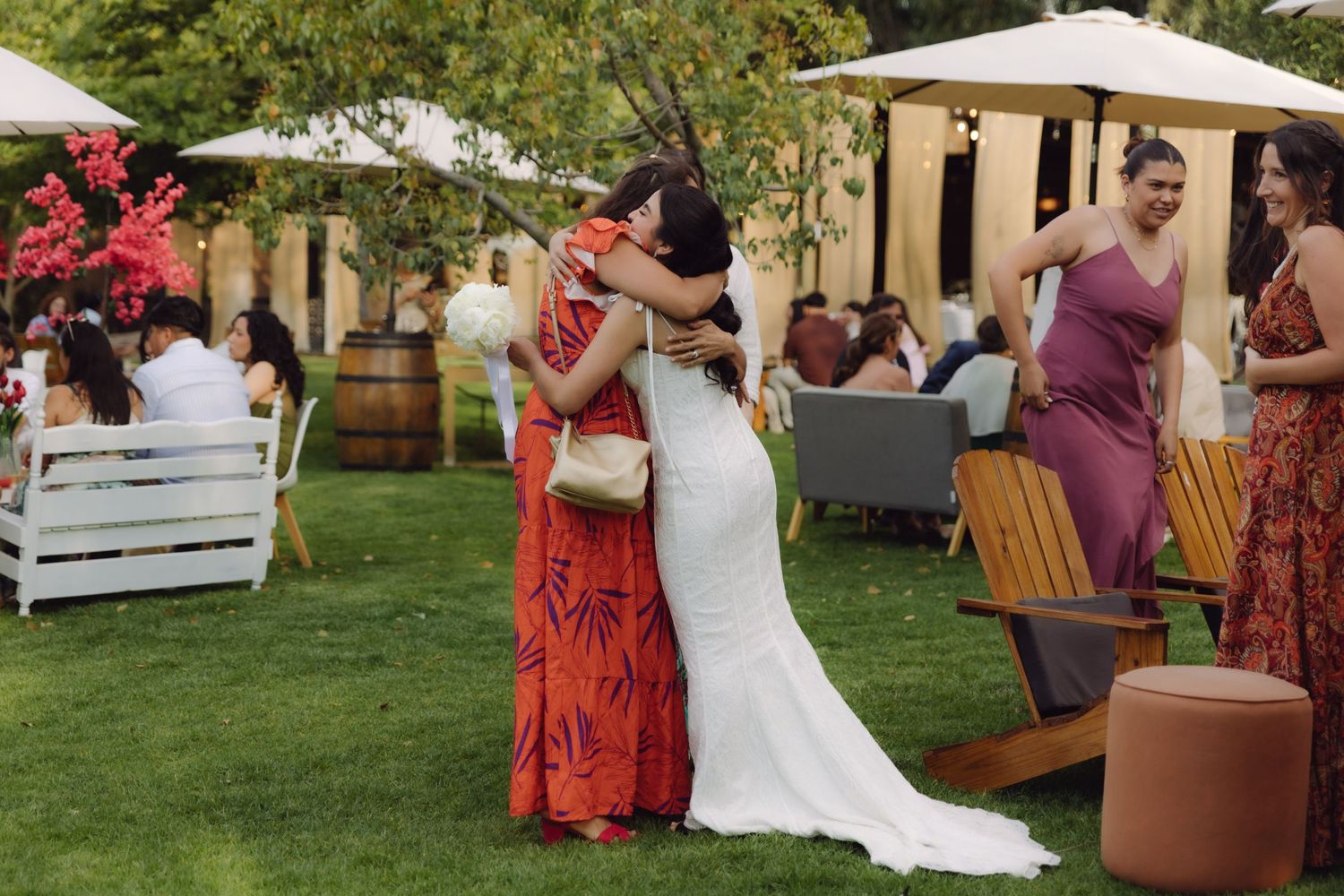 Bride in white gown embracing a guest in red floral dress at an outdoor wedding reception on green lawn.