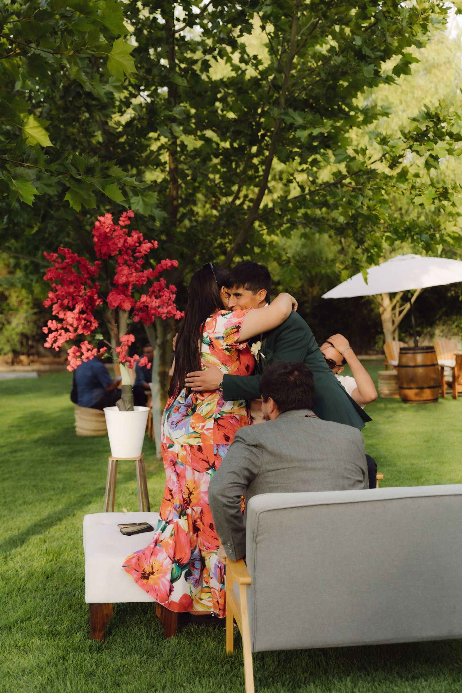 Guest in colorful floral dress sharing a warm hug with a man in green suit at outdoor wedding.