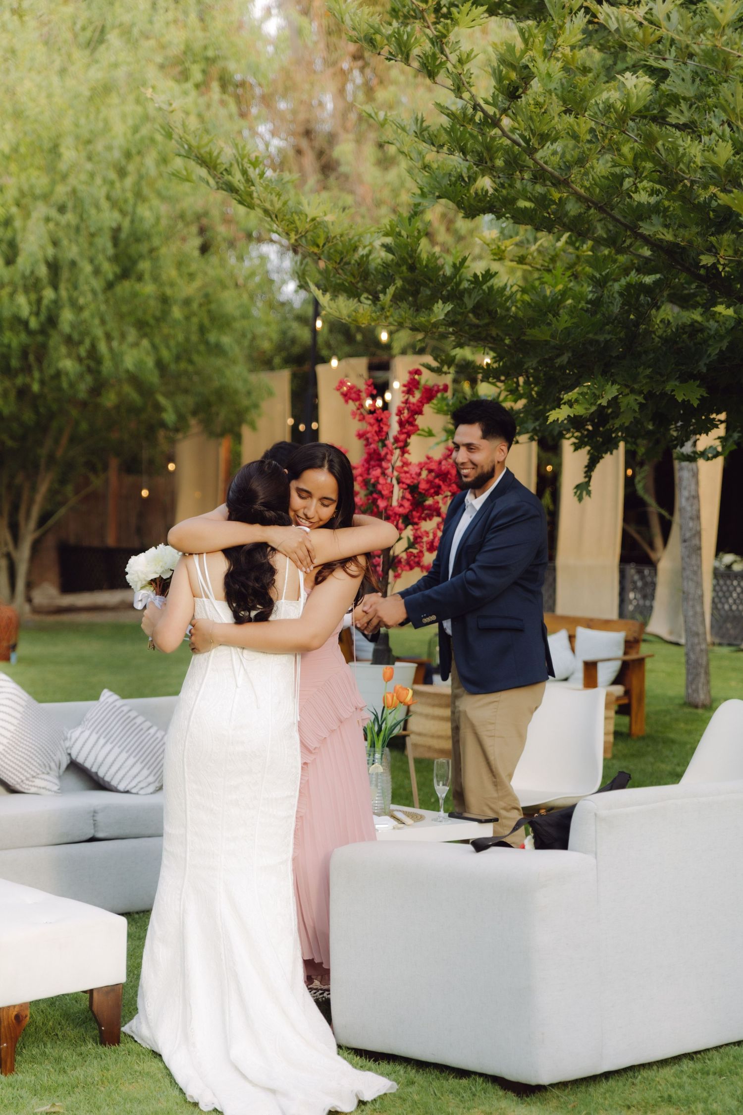 Bride in white gown hugging bridesmaid in pink dress while groom in blue suit watches at outdoor wedding.
