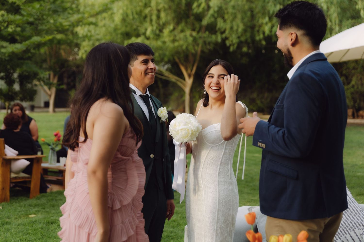 Smiling bride holding white bouquet chatting with guests in pink dress and blue suit at garden wedding.