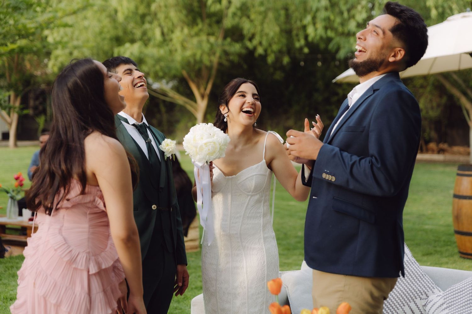 Bride in white dress laughing joyfully with wedding guests including groom and woman in pink at outdoor reception.