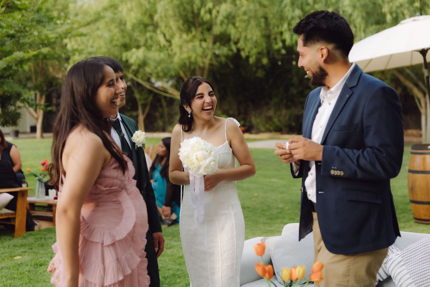 Bride in white gown laughing with bridesmaid in pink and male guest in blue suit at outdoor wedding.