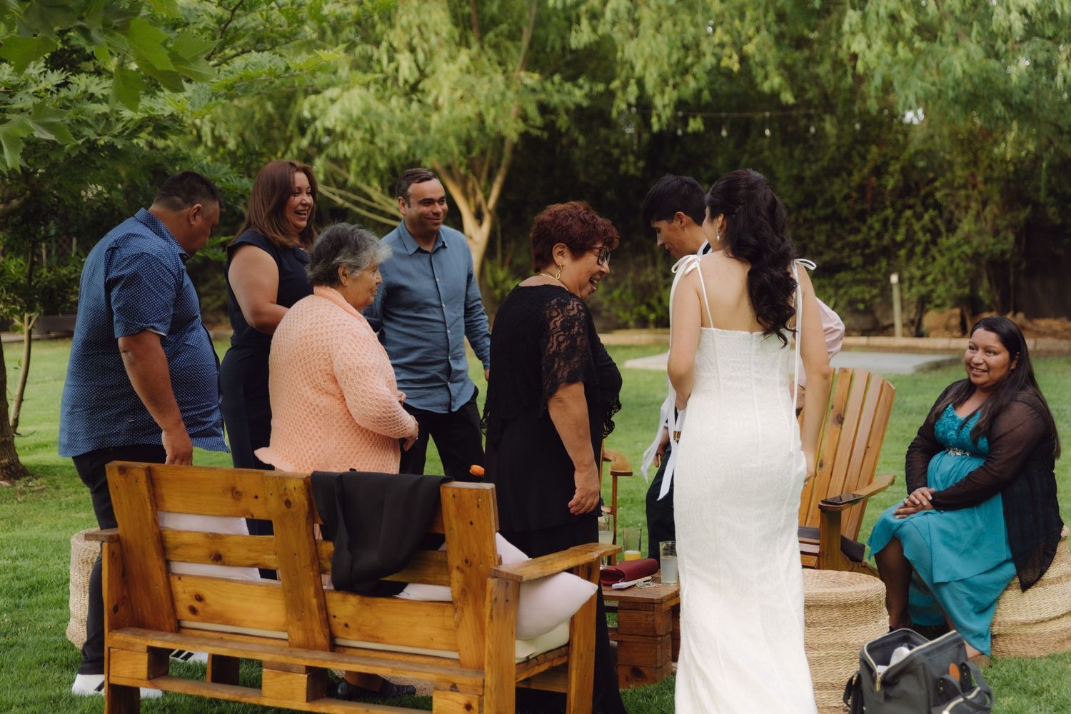Bride in white wedding dress greeting elderly seated guests at a rustic outdoor garden wedding reception.