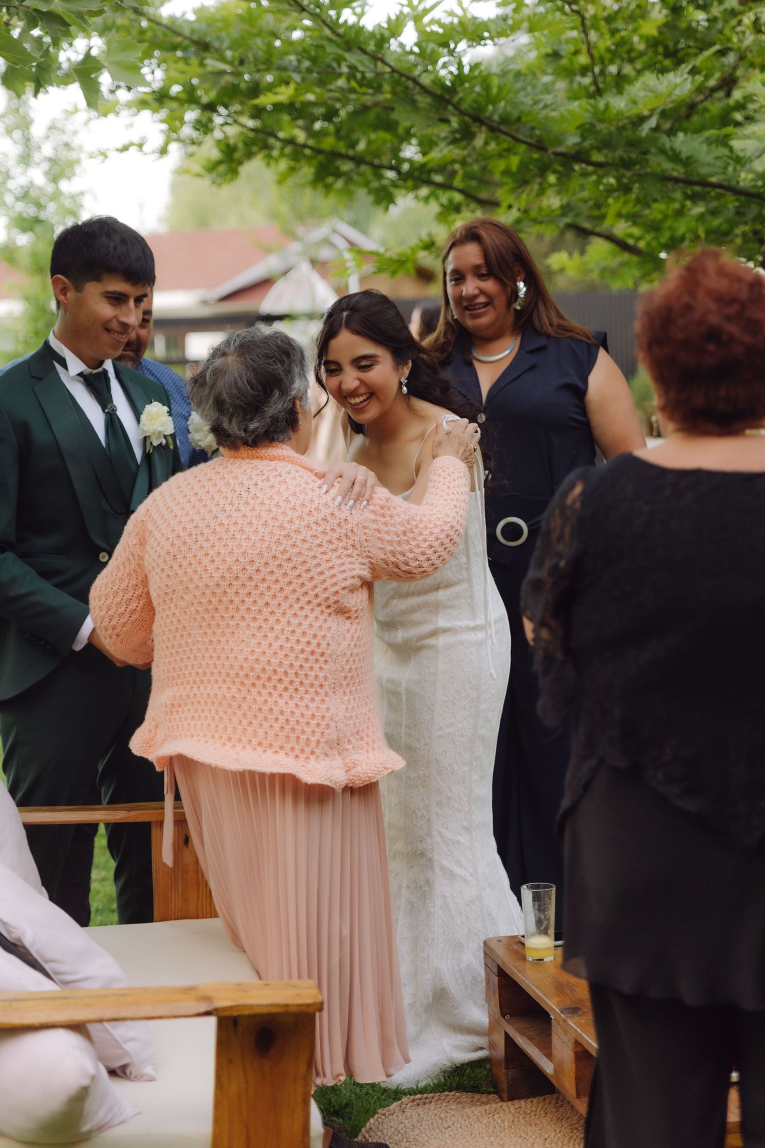 Bride in white gown smiling warmly as elderly woman in pink sweater embraces her at outdoor wedding.