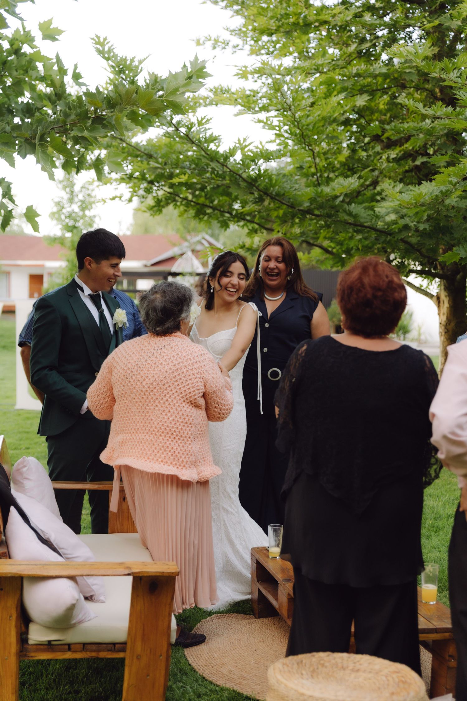 Bride in white dress smiling with groom in green suit and guests under lush green tree at wedding.