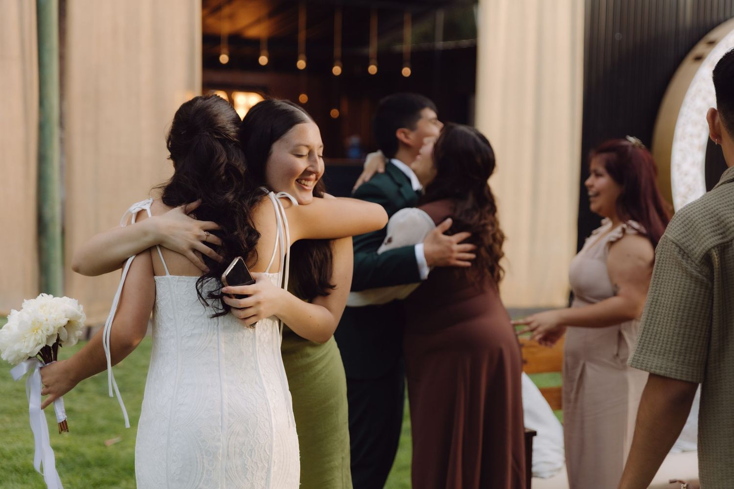 Bride in white dress laughing while hugging wedding guests outdoors at elegant evening reception.