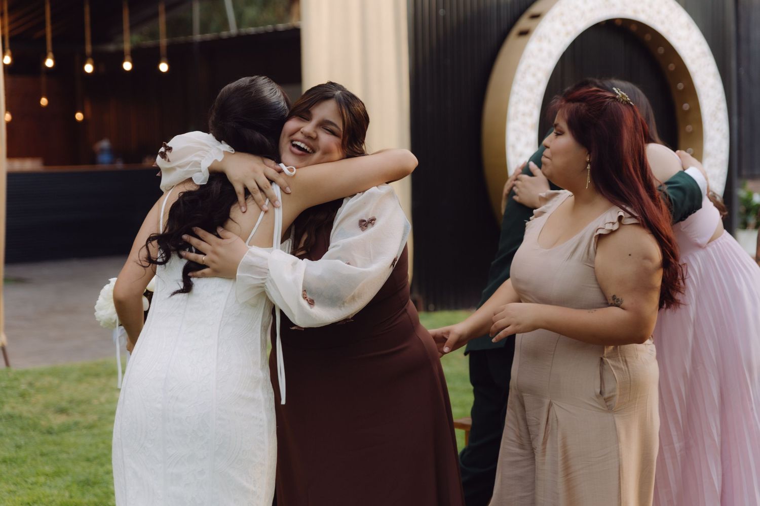 Joyful bride in white gown embracing smiling guest in brown dress at outdoor wedding celebration.