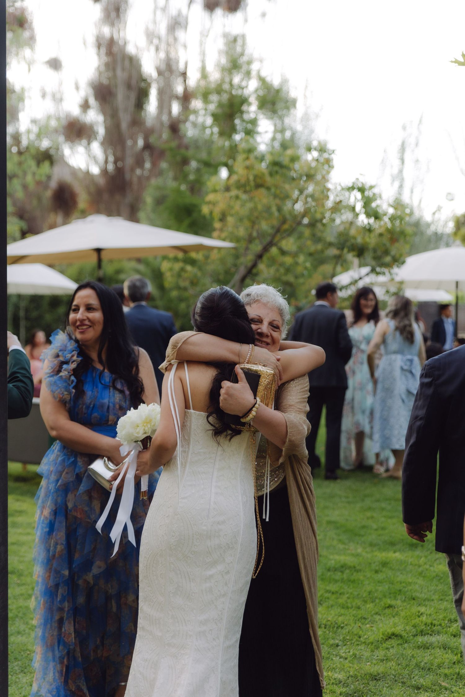 Bride in white dress receiving warm hug from elderly woman at outdoor garden wedding reception.
