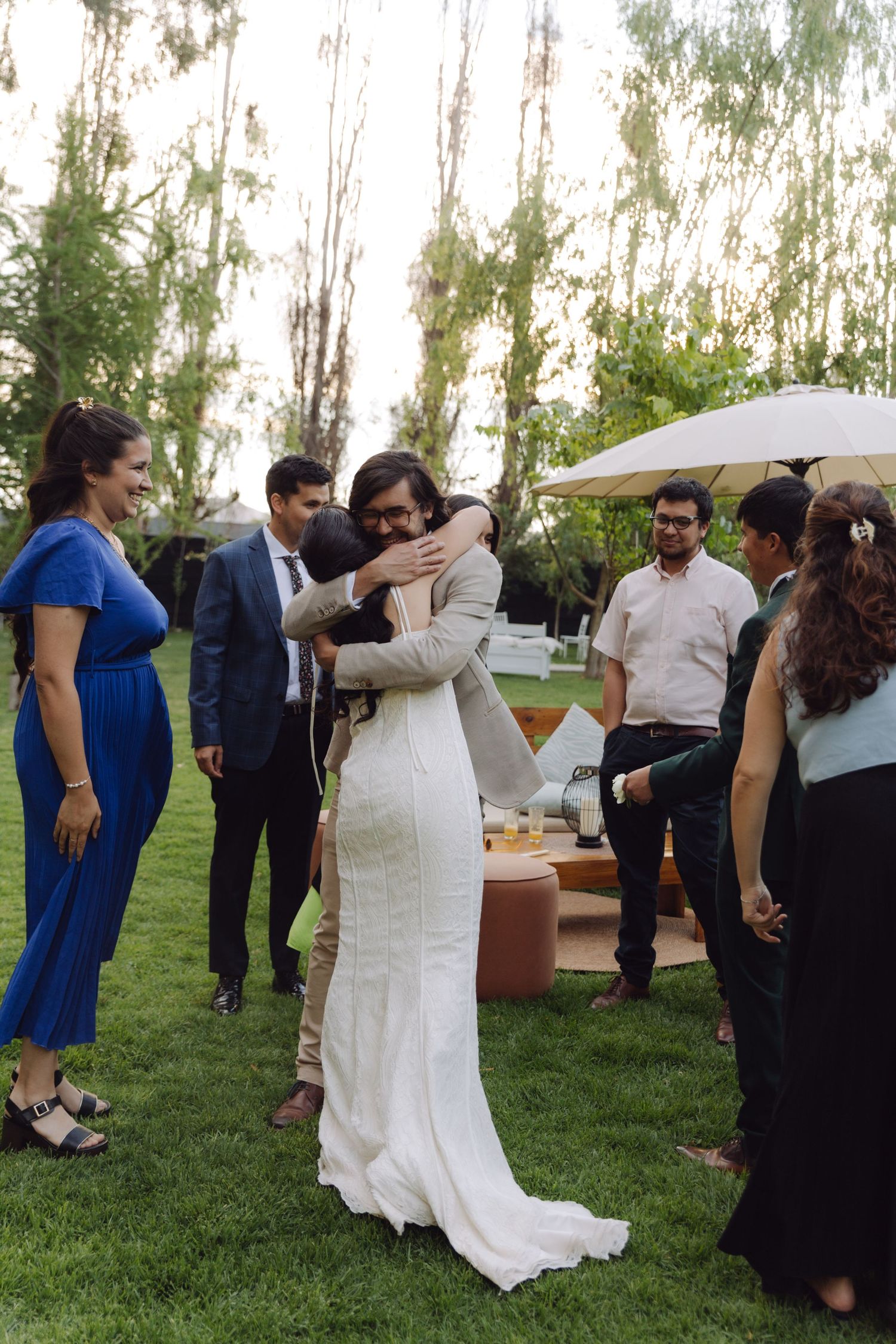 Bride in white gown hugging male guest on green lawn surrounded by wedding party outdoors.
