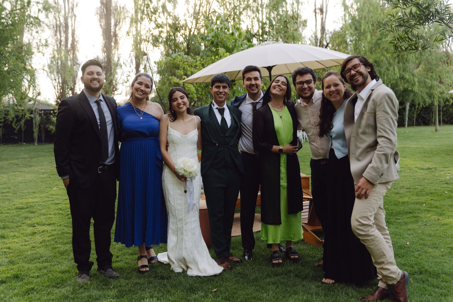 Wedding party group photo with bride and groom surrounded by friends on lush garden lawn.