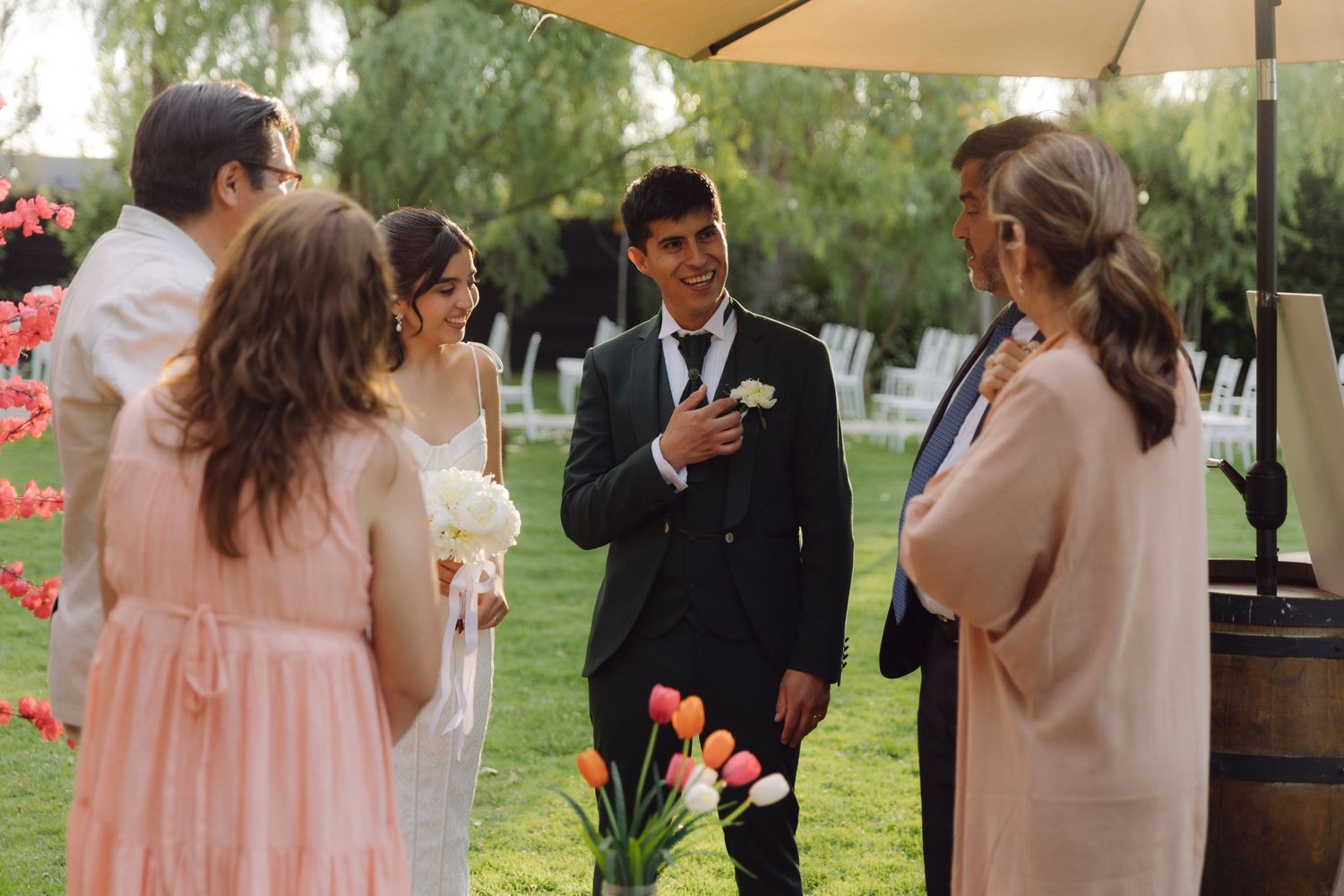 Groom in dark suit greeting wedding guests near colorful tulip arrangement at outdoor ceremony.