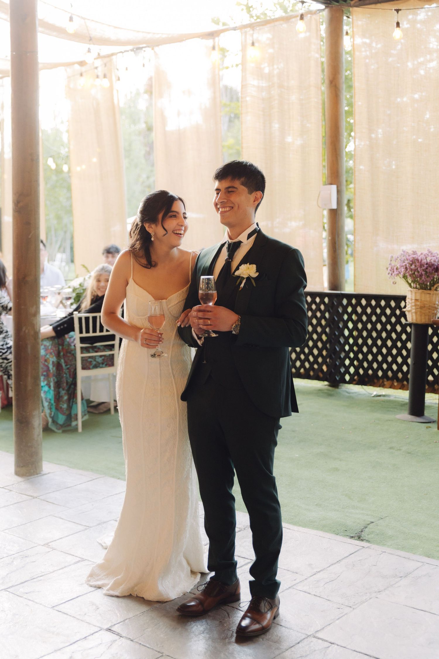 Bride and groom smiling and holding champagne glasses under string lights at wedding reception.