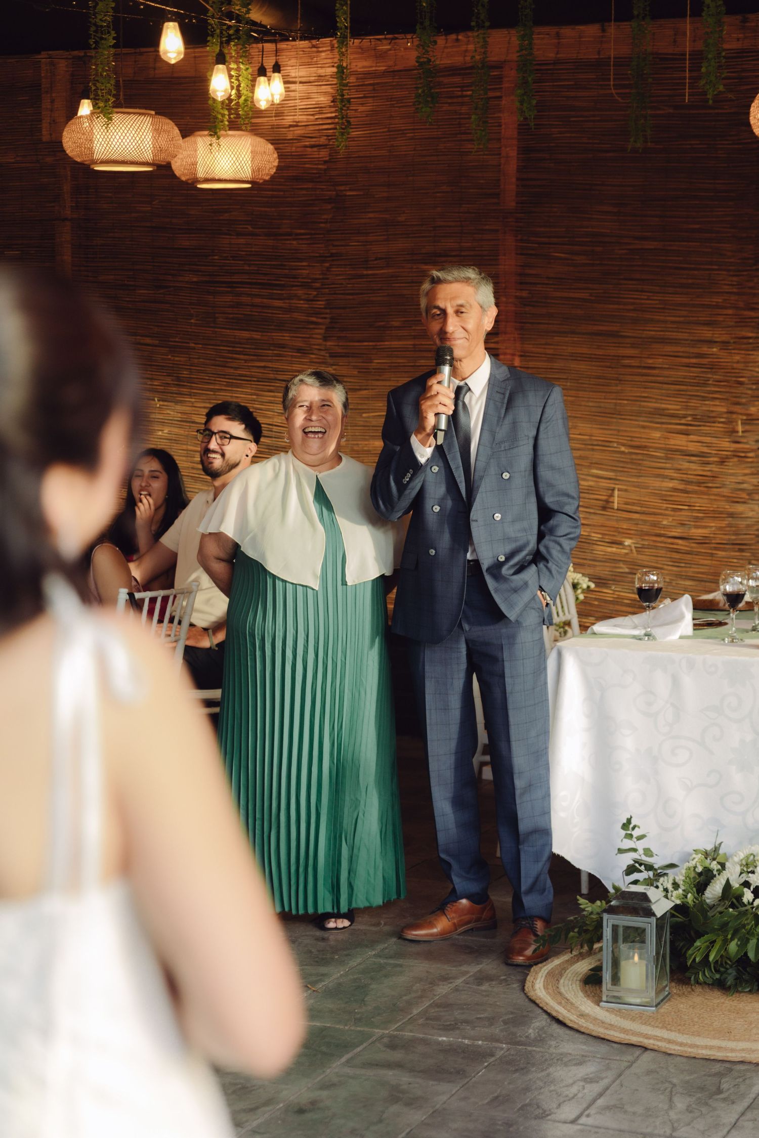 Older man in blue suit giving speech with microphone at indoor wedding reception dinner.