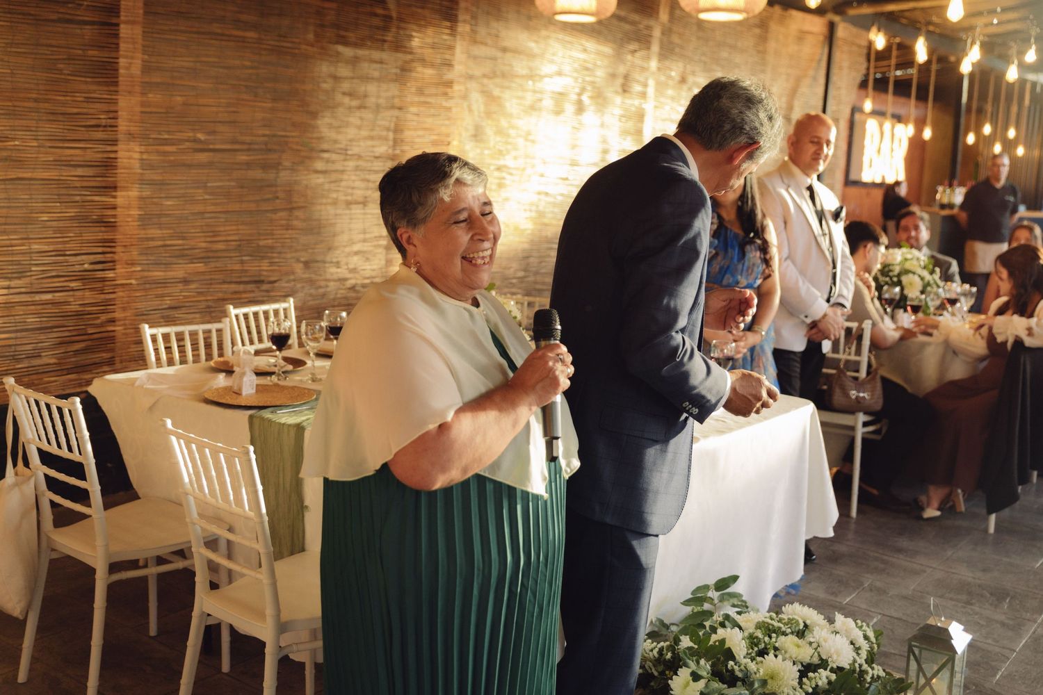 Older woman in green pleated dress speaking into microphone at elegant indoor wedding reception.