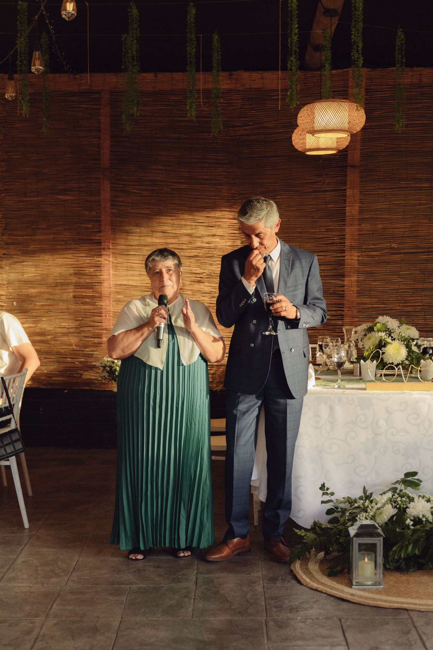 Couple in formal attire giving emotional wedding toast with microphone at indoor reception dinner.
