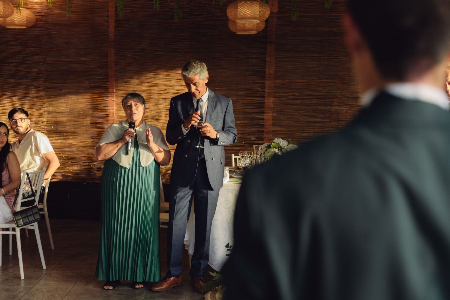 Couple giving wedding speech to guests in warmly lit reception venue with wicker pendant lights.
