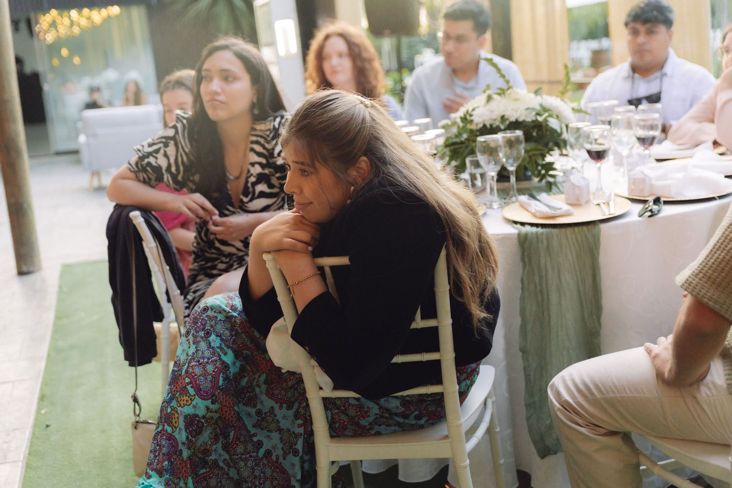 Wedding guests seated at outdoor reception table with green floral centerpiece in natural light.