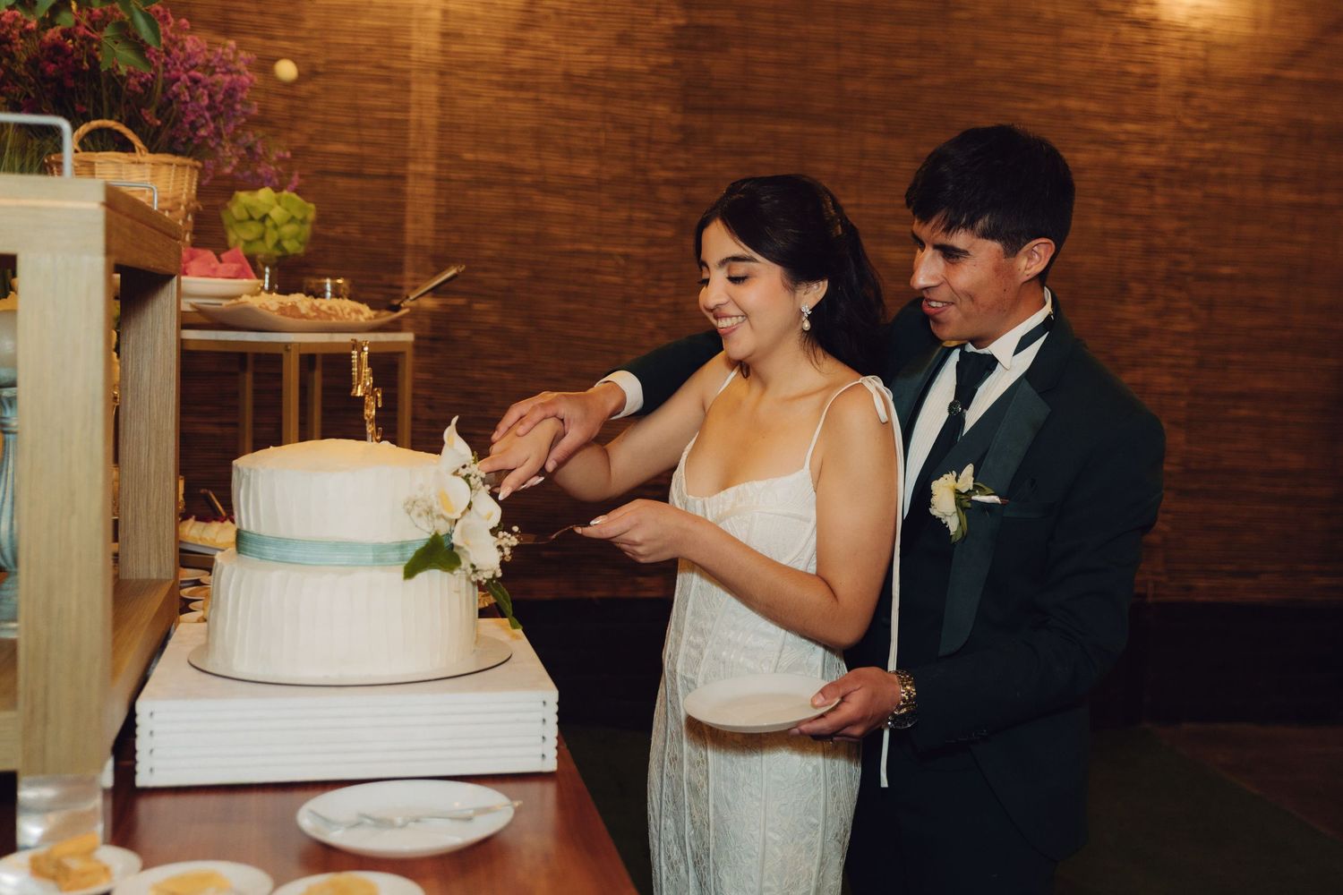 Bride and groom smiling while cutting white wedding cake decorated with flowers together.