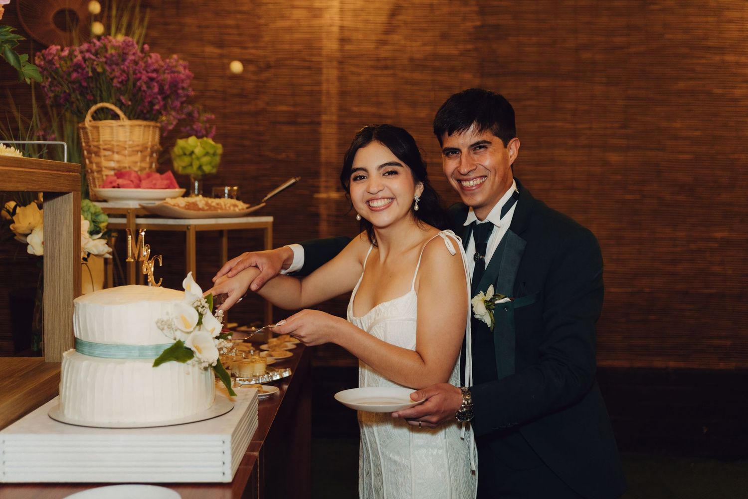 Smiling bride and groom posing together while cutting their white floral wedding cake.
